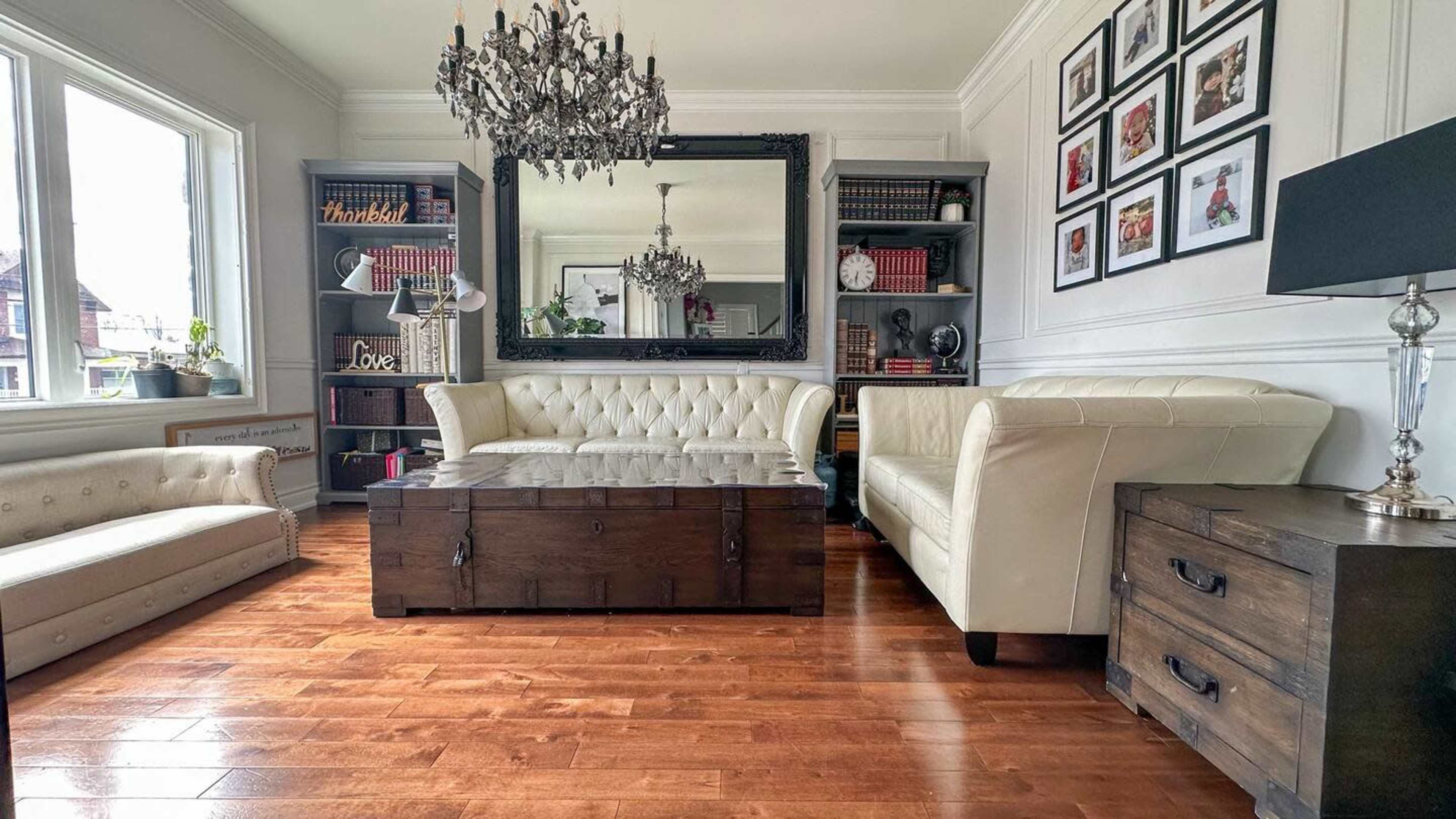 A well-furnished living room with a chandelier, a large mirror, two sofas, a wooden coffee table, and a bookshelf against the wall.