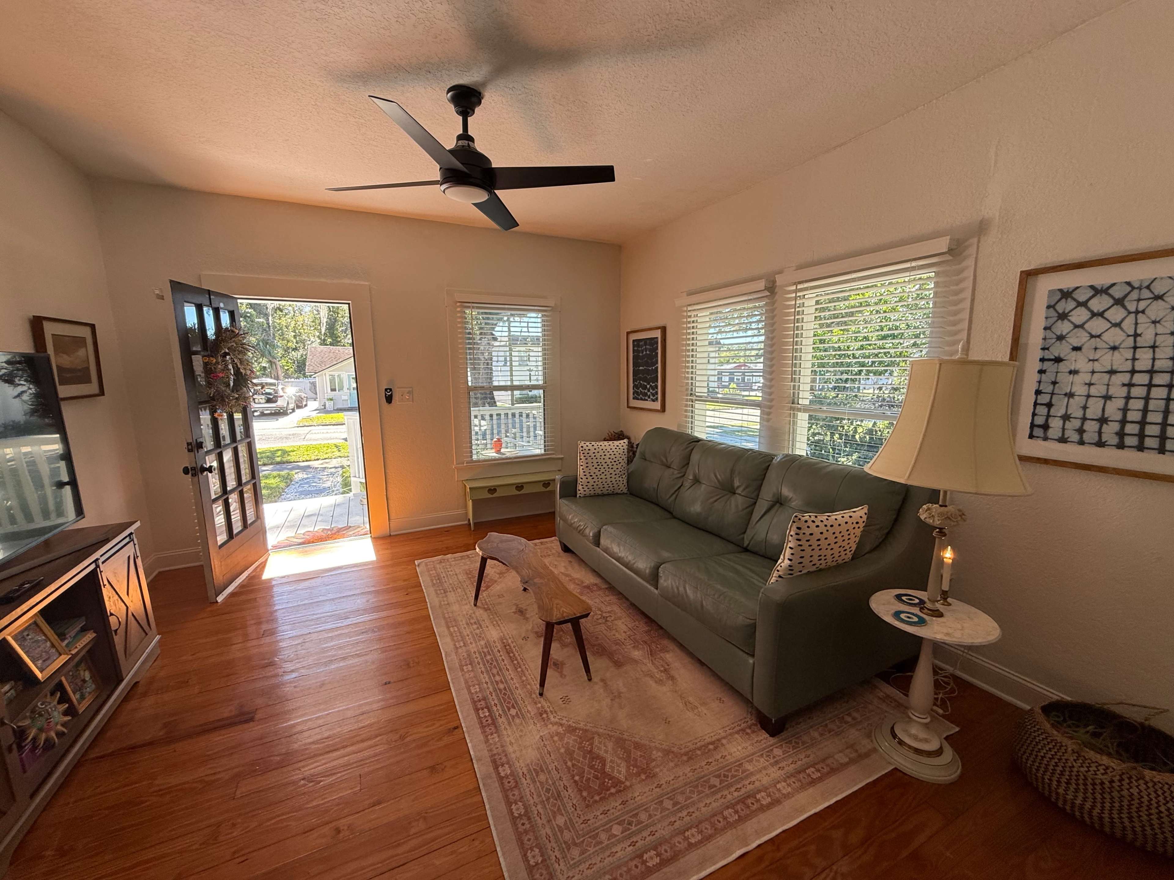 A light-filled living room features a green sofa, a wooden coffee table, and large windows, with a view of a porch and outdoor greenery through the open front door.