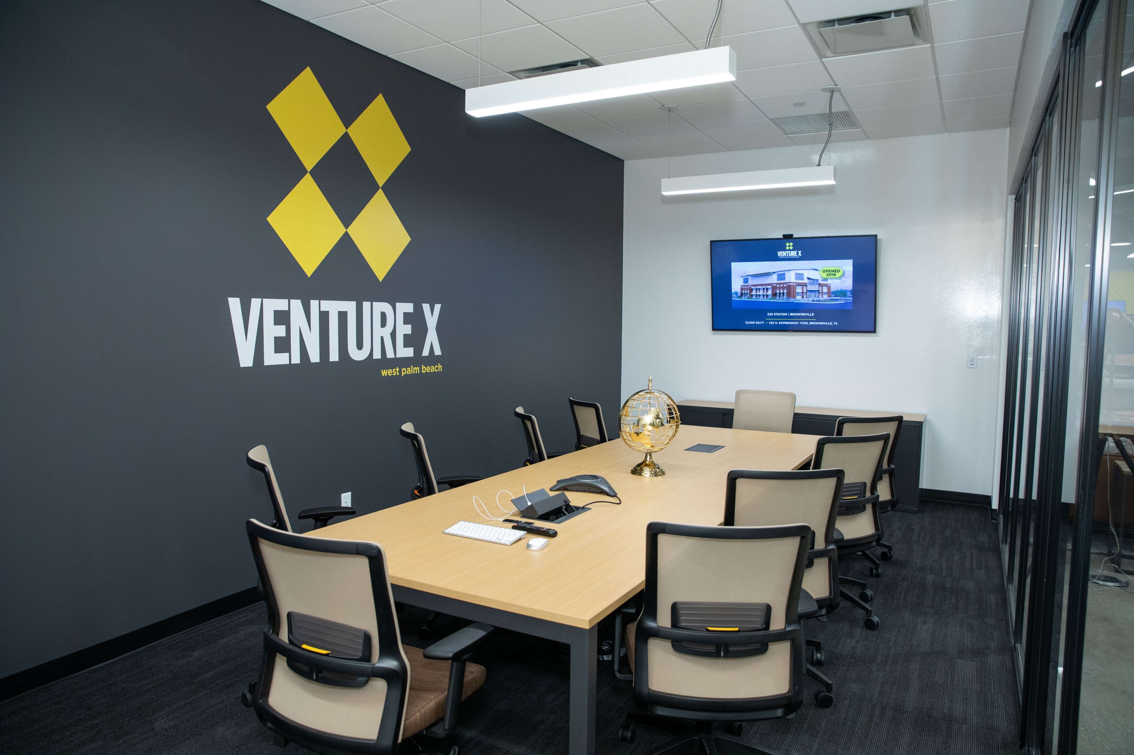 The image shows a modern conference room with a large table, several chairs, a wall-mounted screen, and a wall featuring the "Venture X" logo.