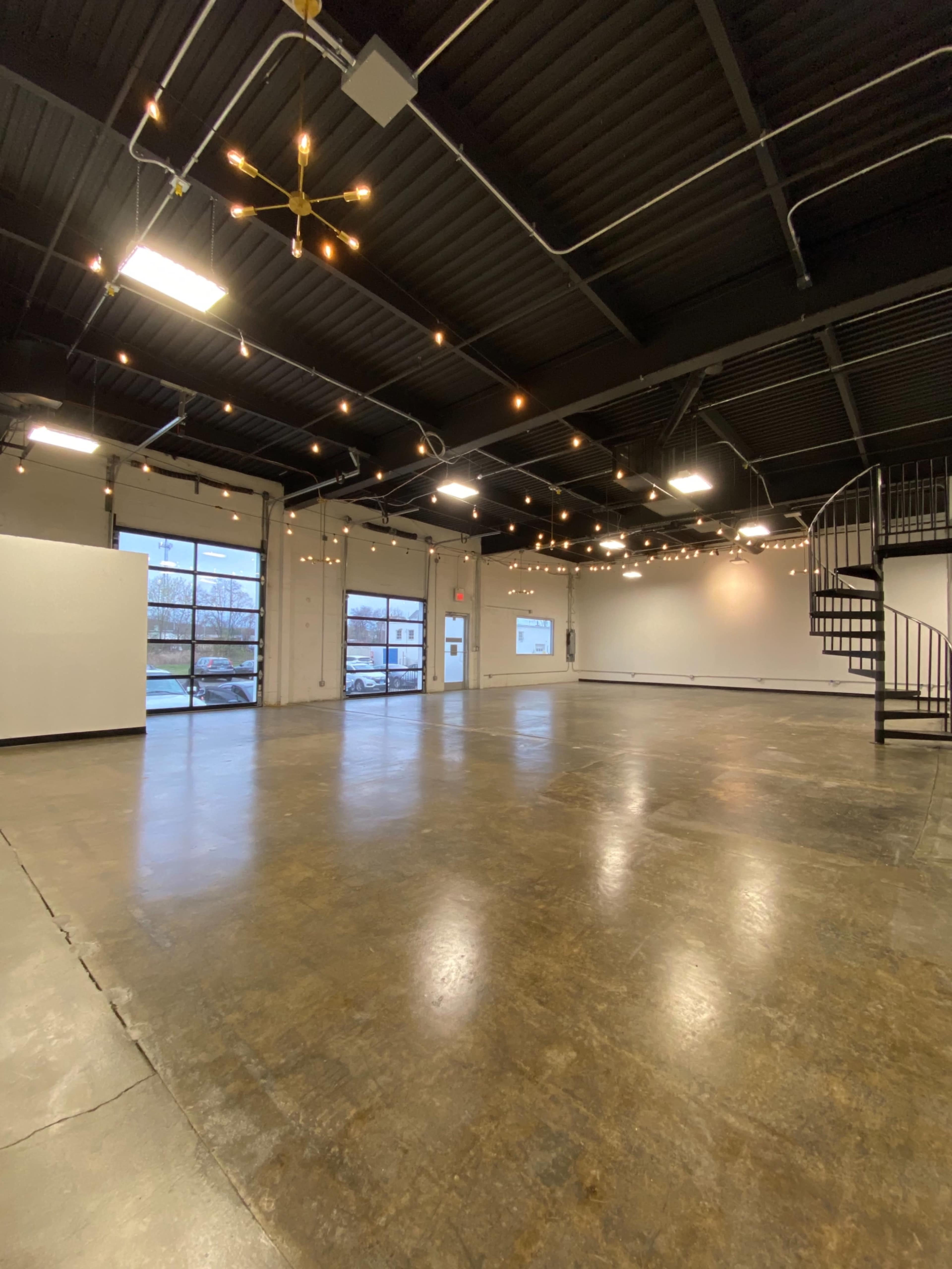 An empty industrial-style space with a polished concrete floor, high ceilings, and hanging string lights, featuring large windows on one side and a spiral staircase in the corner.
