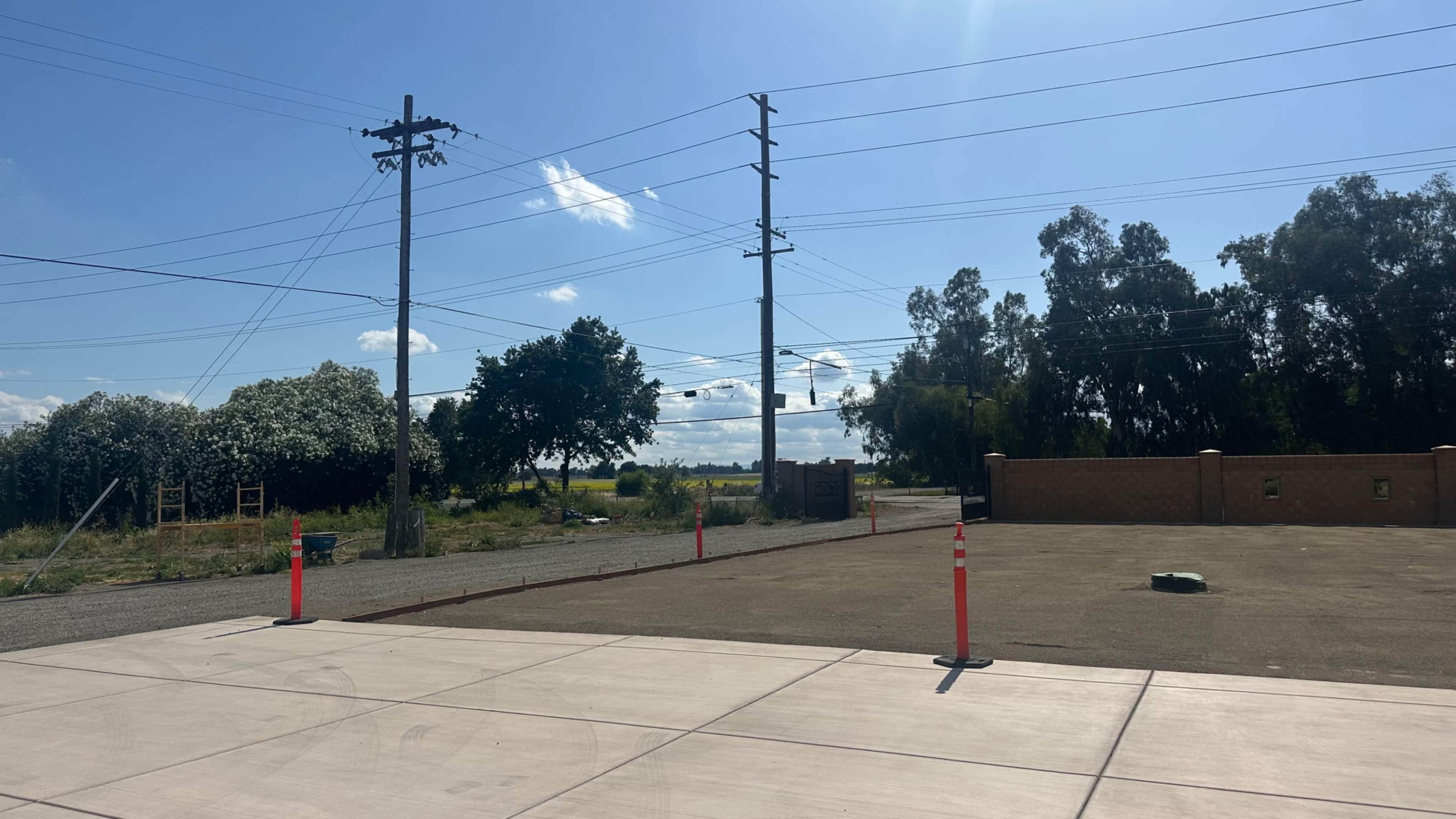 The image shows an empty lot with concrete pavement, utility poles, and a line of trees against a blue sky with scattered clouds.
