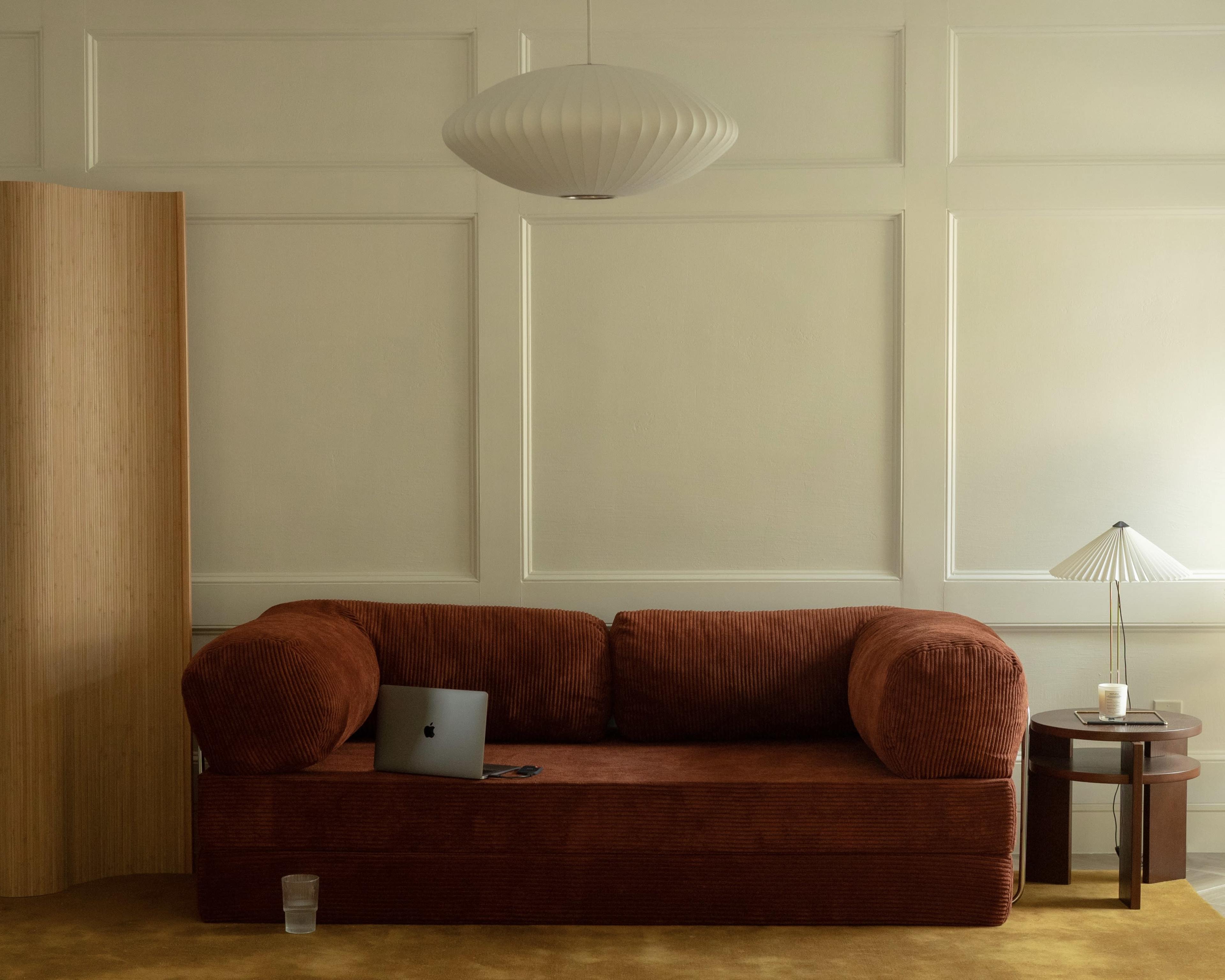 A brown corduroy sofa sits against a wall with paneling, accompanied by a small round table with a lamp and a glass of water nearby.