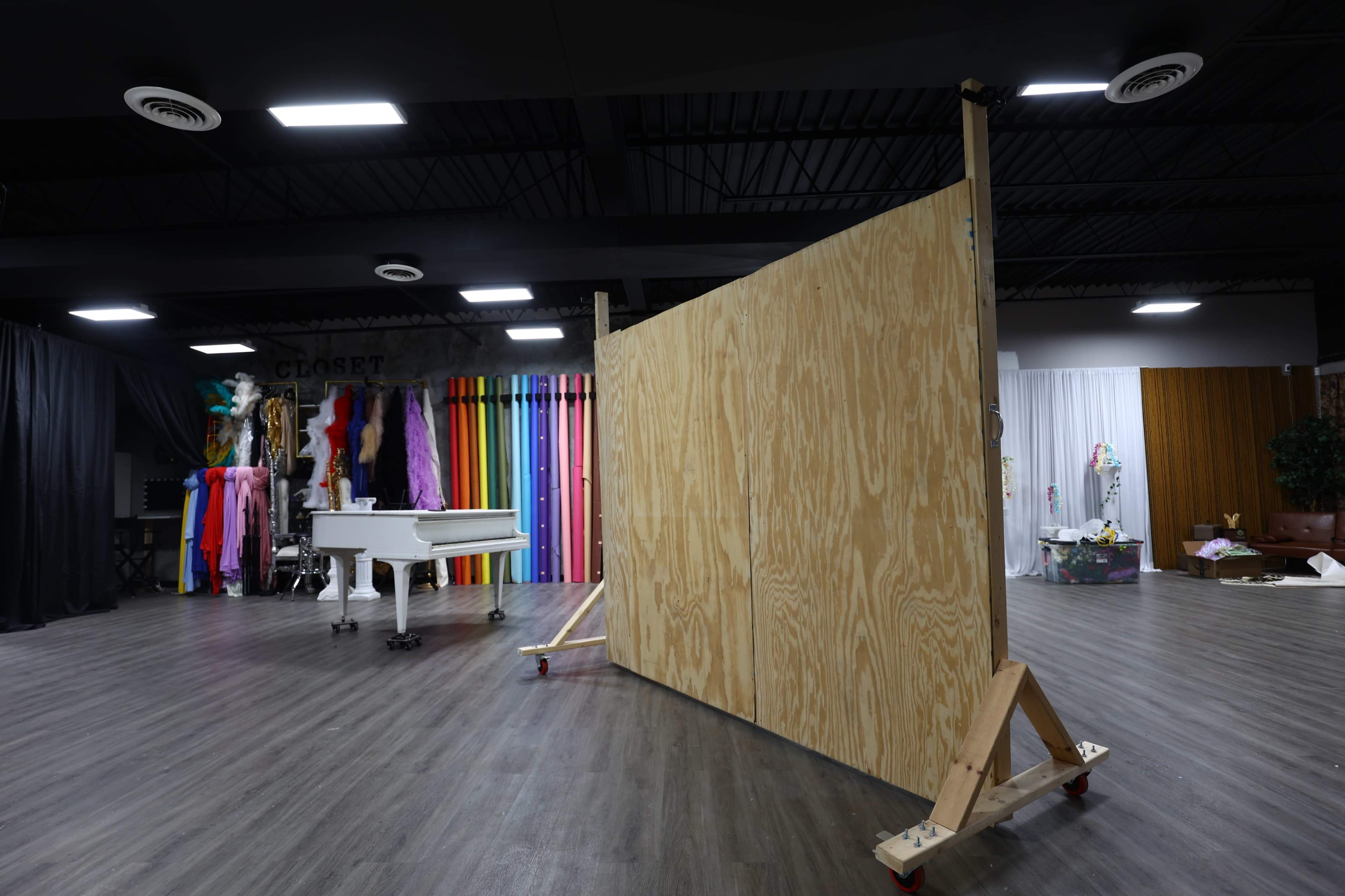 The image shows a spacious studio with a wooden partition on wheels, a white piano, and colorful backdrops arranged along one wall.