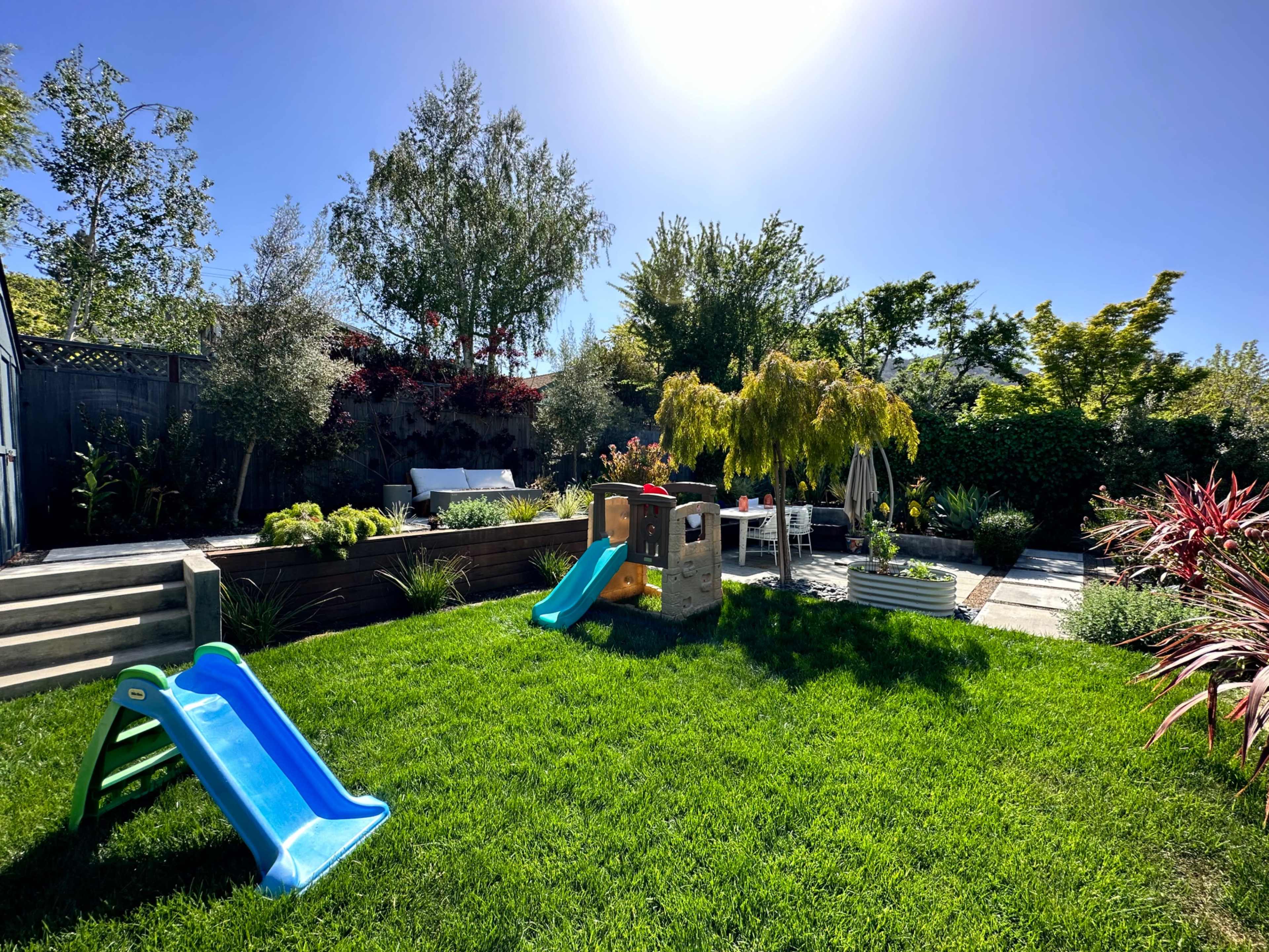 A sunny backyard features green grass, a small slide, a playhouse, and various plants and seating areas.