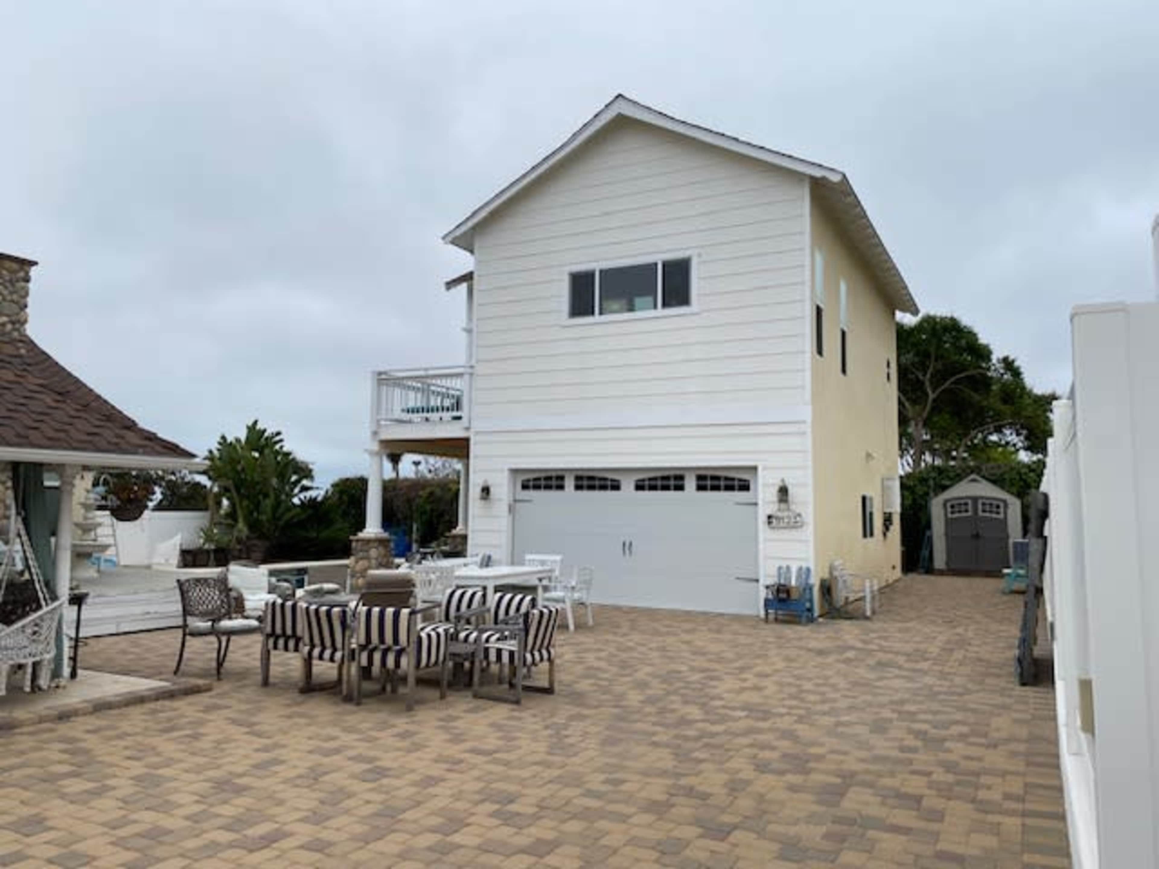 A two-story house with a white exterior and a garage is situated on a paved patio area surrounded by outdoor furniture.