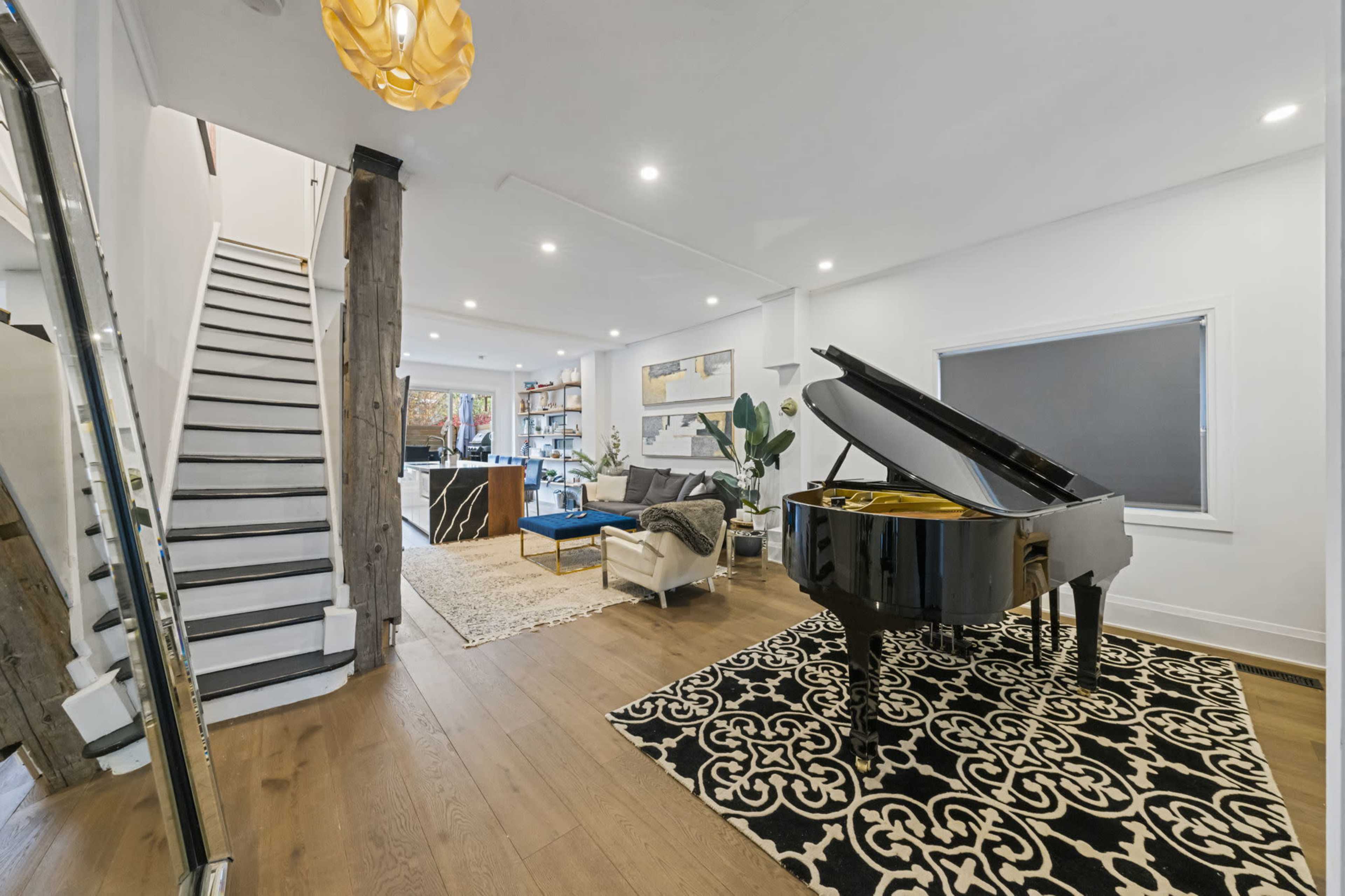 A modern living space featuring a grand piano, stairs leading to an upper level, and a patterned rug on wooden flooring.