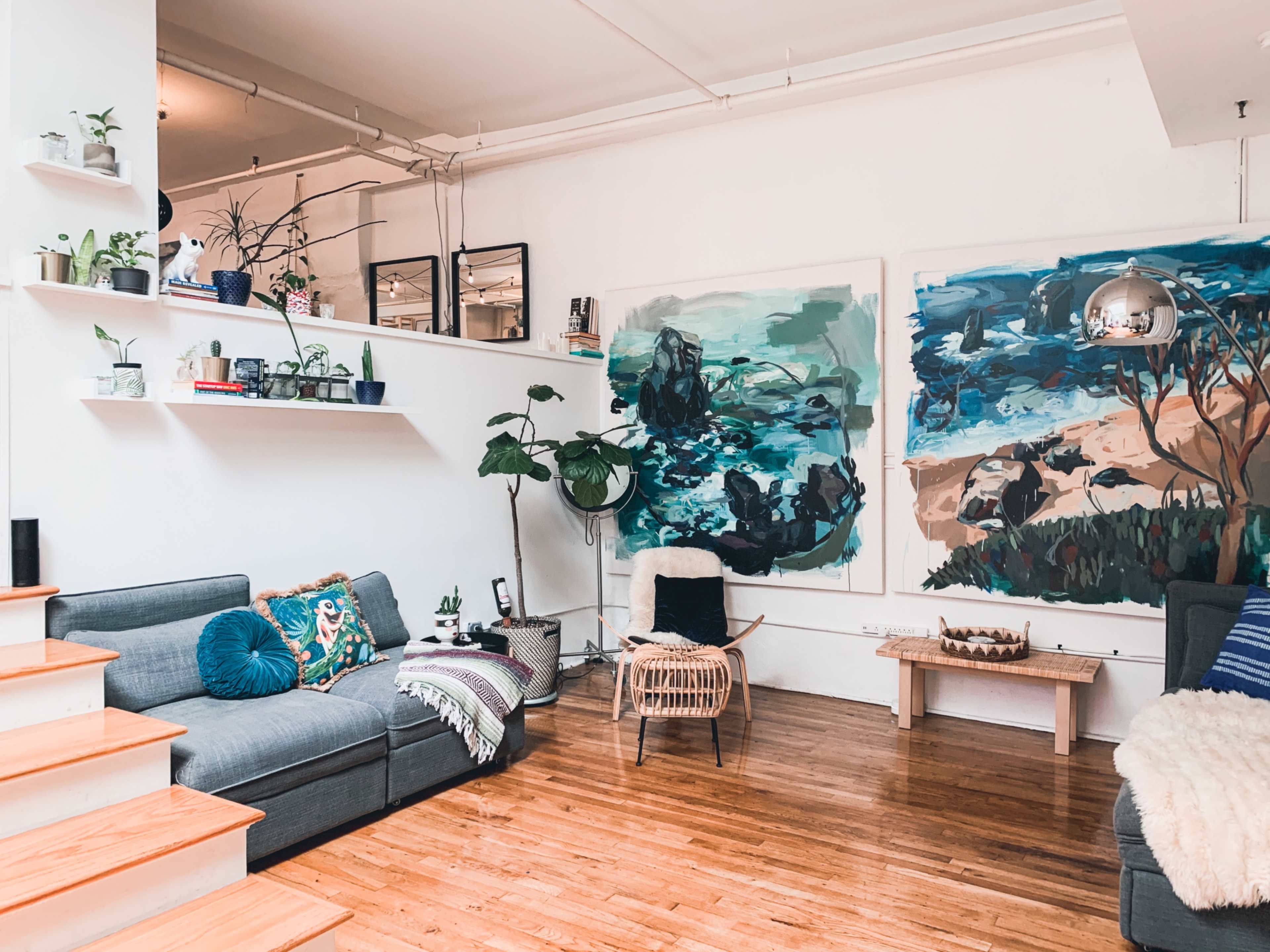 A bright living space features a cozy seating area with two sofas, large abstract paintings on the wall, and plants arranged on shelves and the floor.