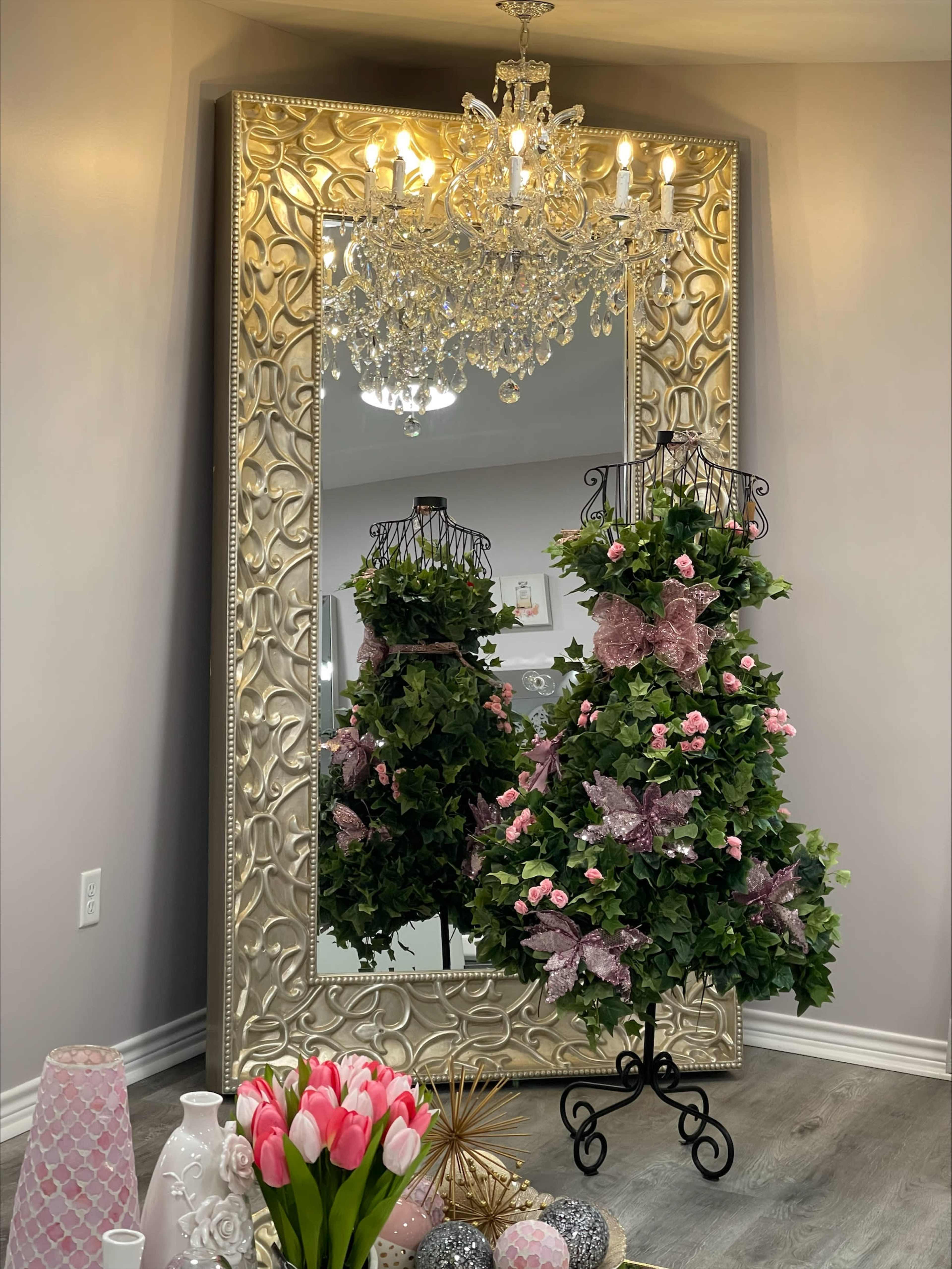 A decorative mirror with an ornate frame reflecting a pair of plants adorned with pink bows and flowers, alongside a chandelier and decorative vases.