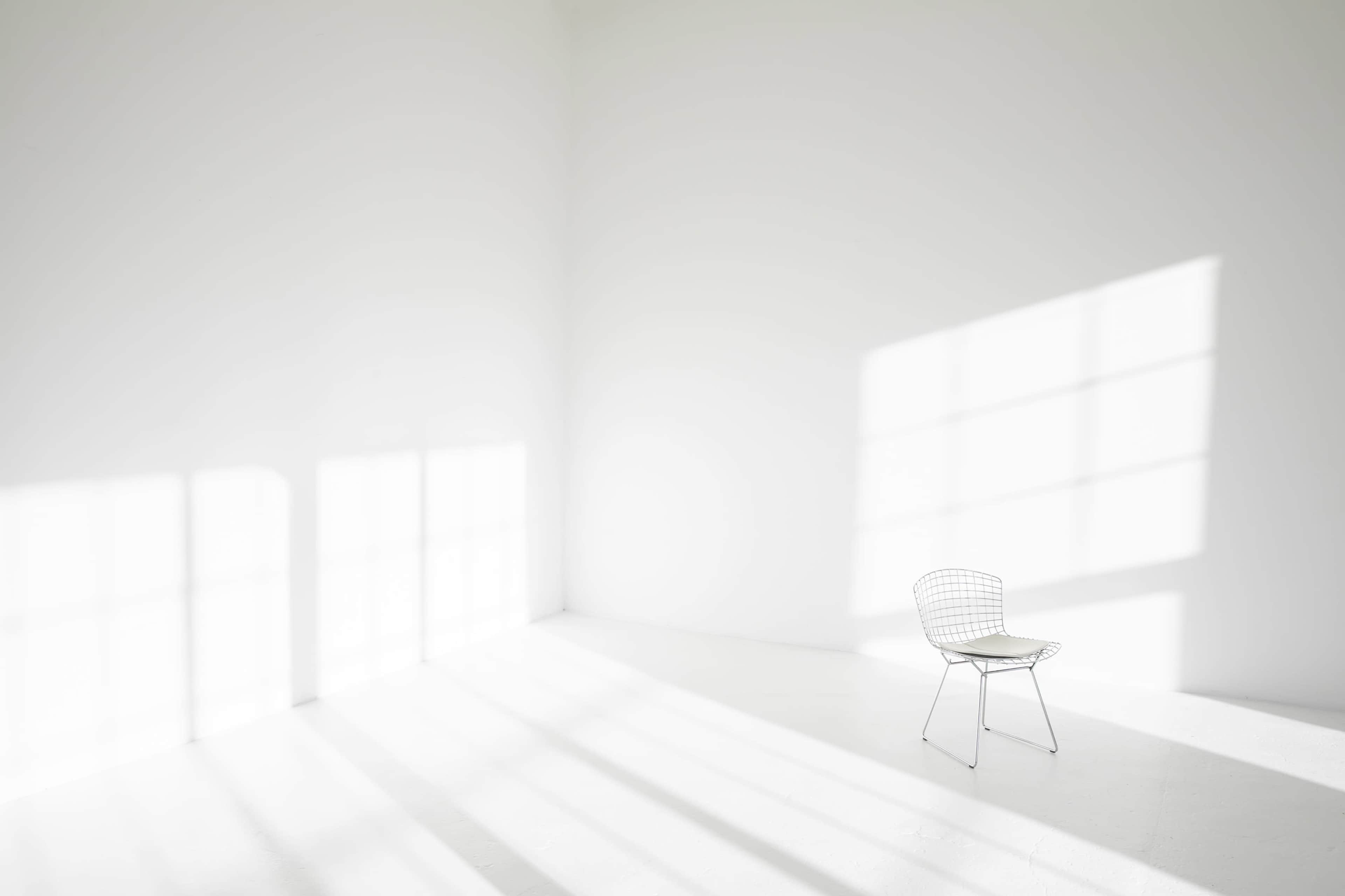 A single wire chair is positioned in a bright, empty room with white walls and long shadows cast by sunlight filtering through large windows.