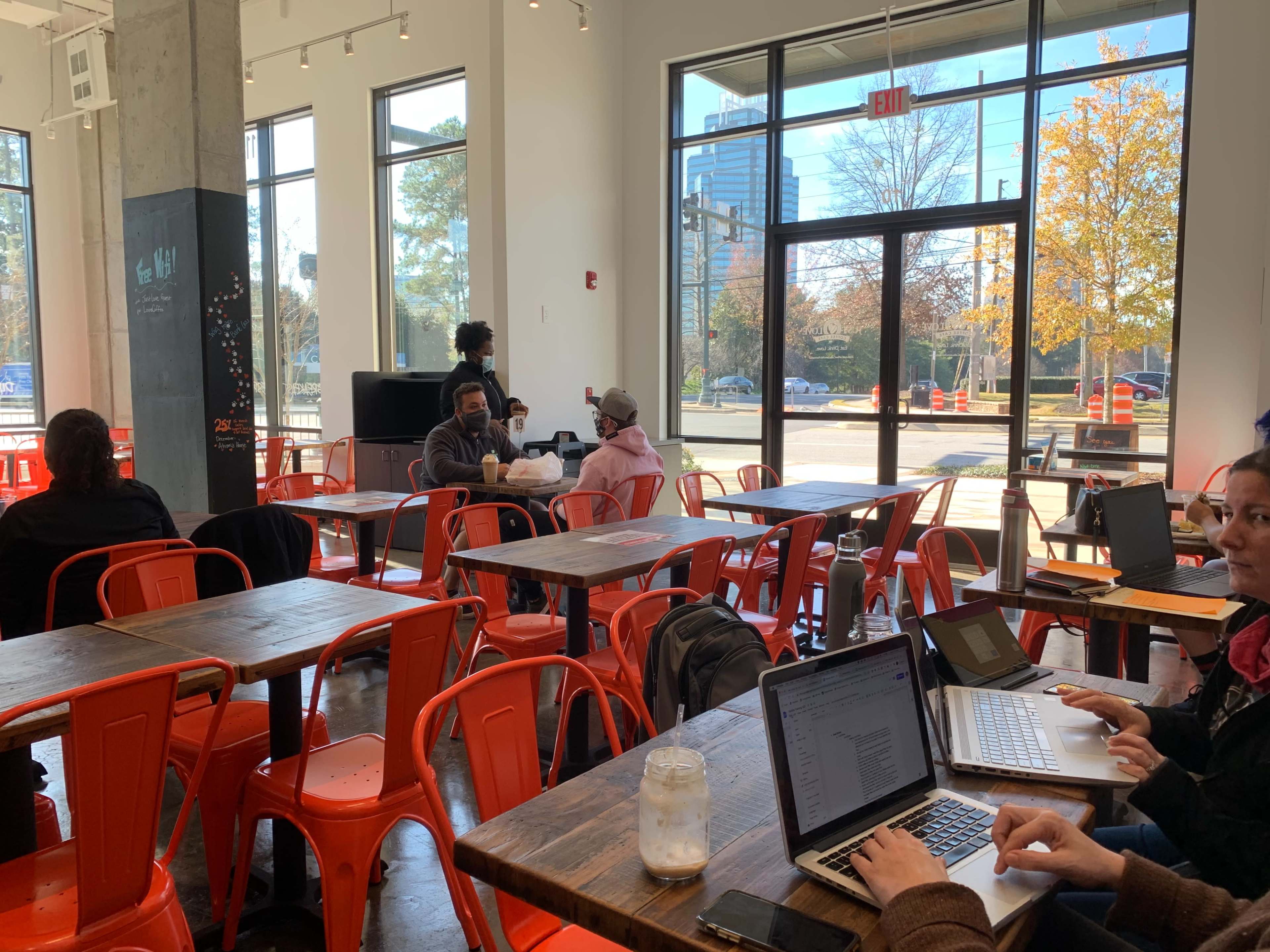 A bright café features several patrons working on laptops at wooden tables with red metal chairs, while others converse in the background near large windows.