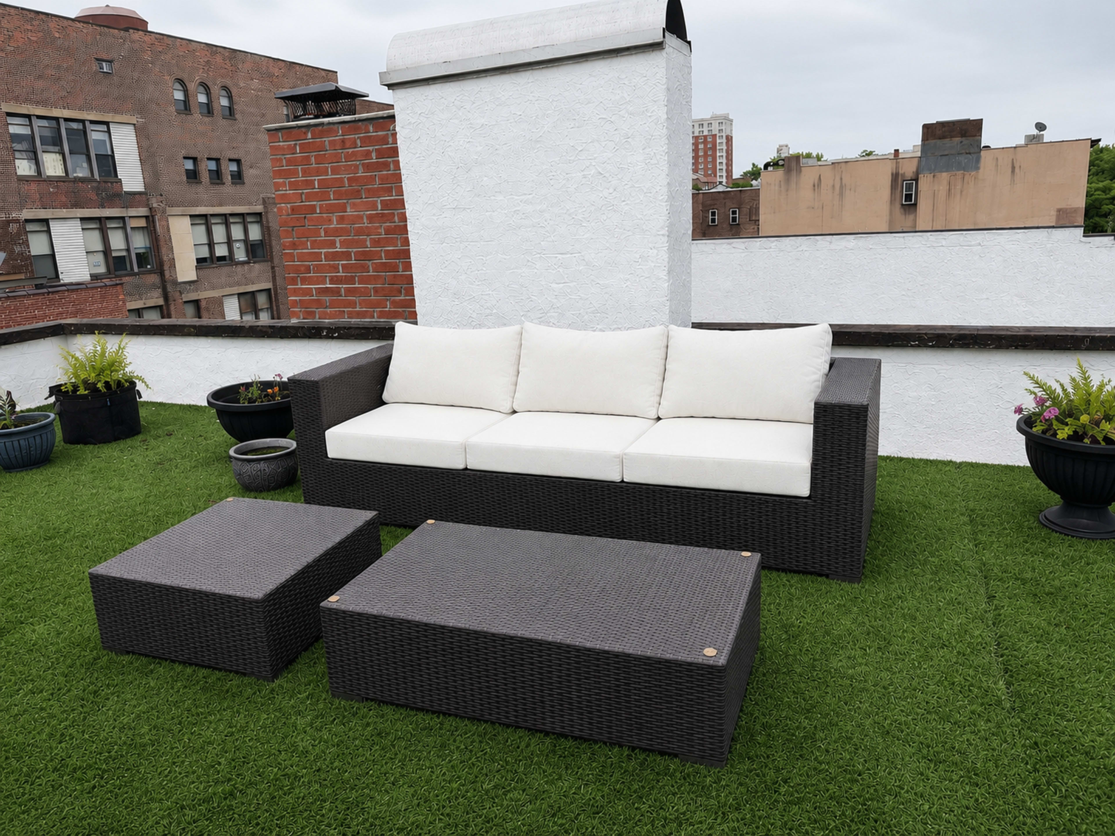 The image shows a rooftop terrace featuring a dark wicker sofa with white cushions, accompanied by two matching wicker coffee tables on artificial grass, surrounded by potted plants.