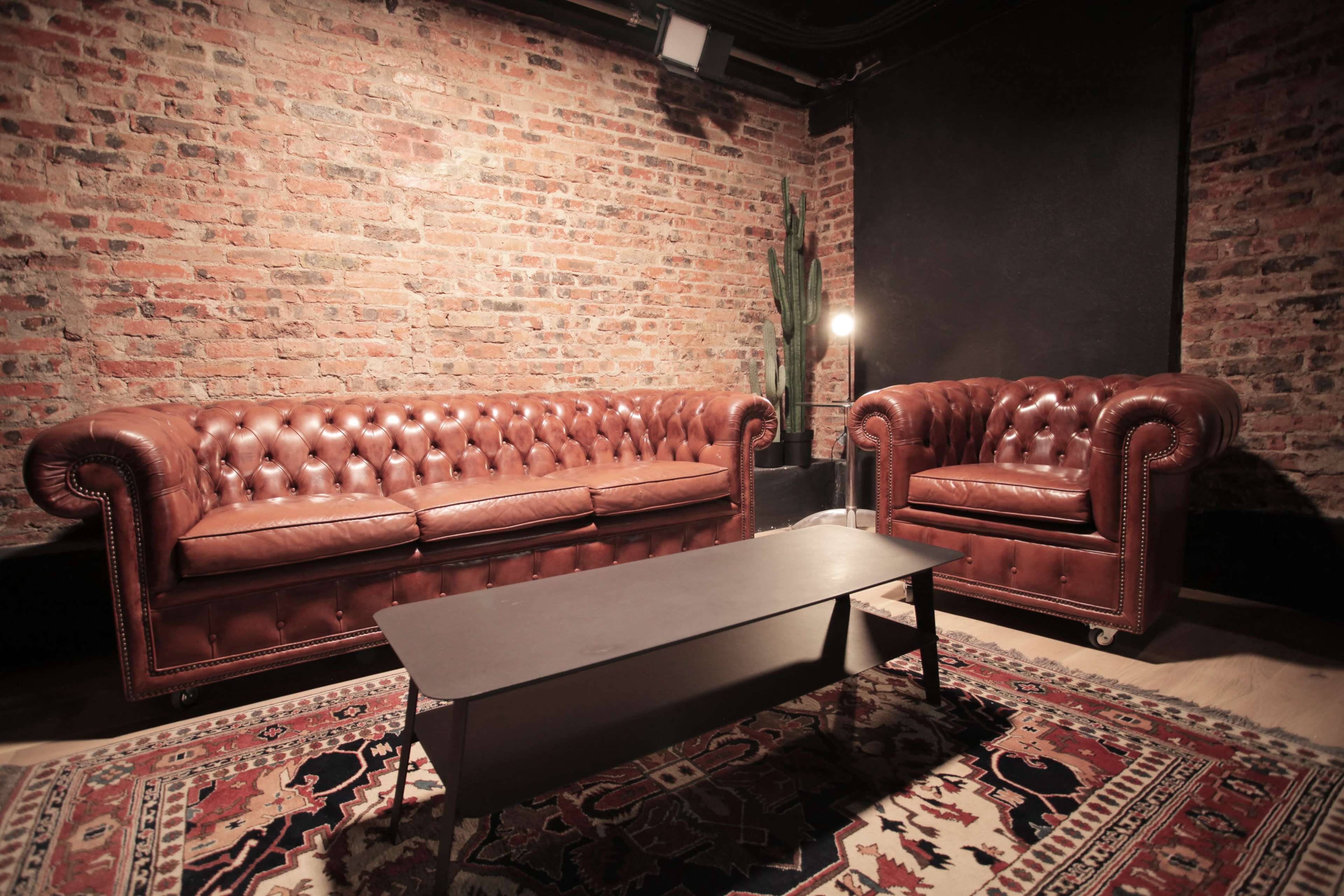 The image features a pair of brown leather Chesterfield sofas positioned next to a low coffee table on a patterned rug, set against a backdrop of exposed brick walls.