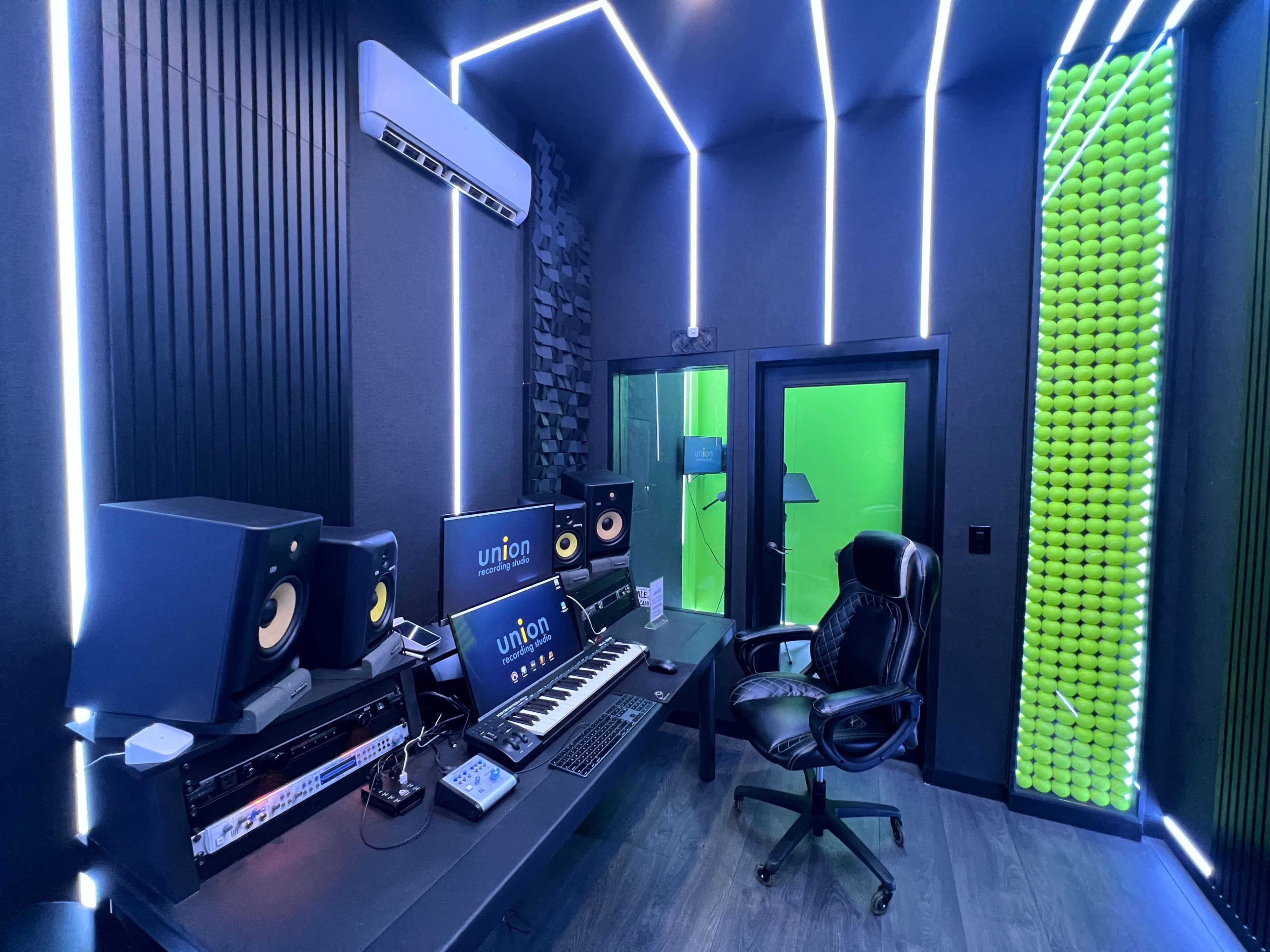 A modern recording studio equipped with a computer, keyboard, speakers, and a black chair, featuring green and blue accent lighting.
