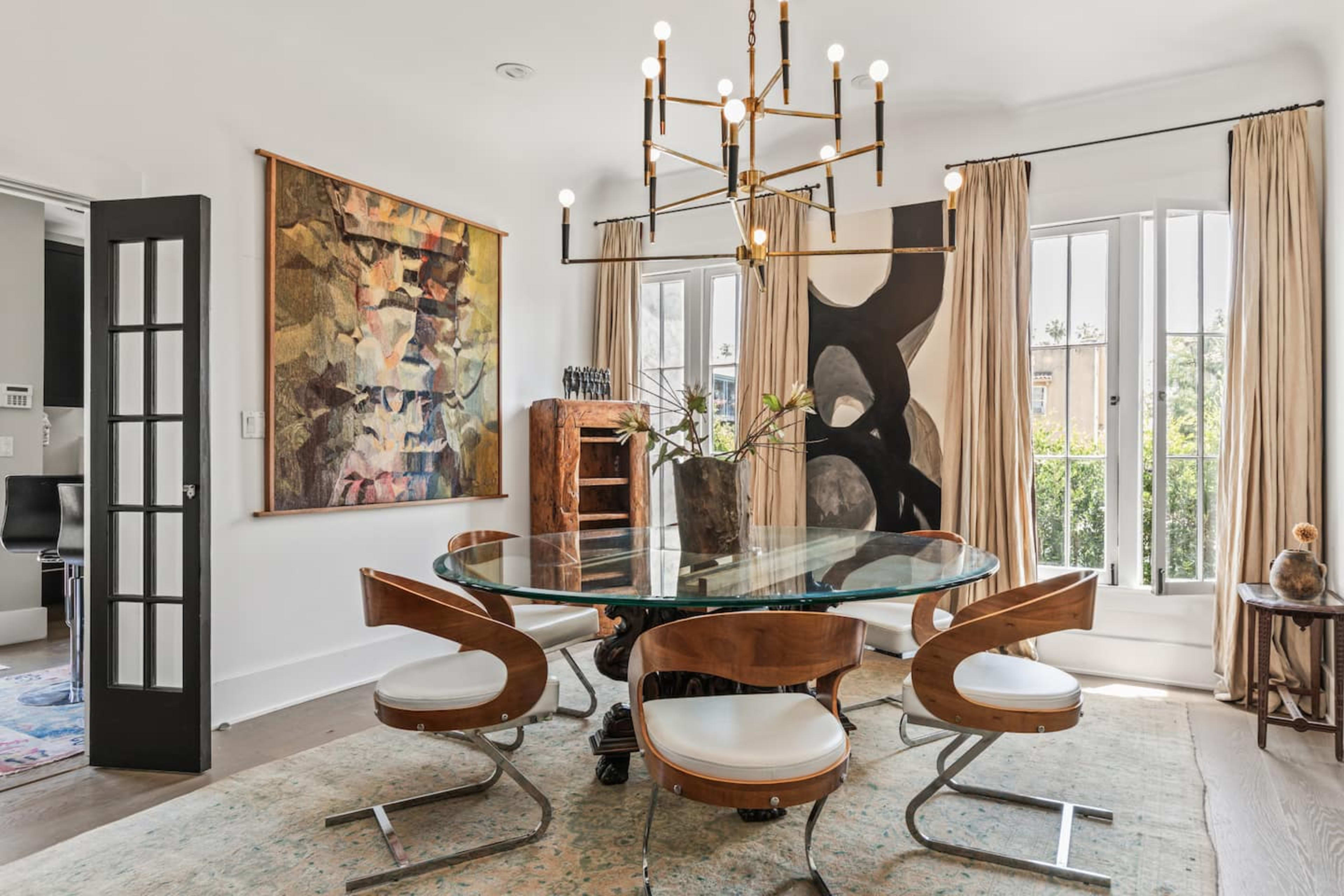 The image shows a modern dining room featuring a round glass table surrounded by stylish chairs, with a large abstract painting on one wall and large windows dressed with light curtains.