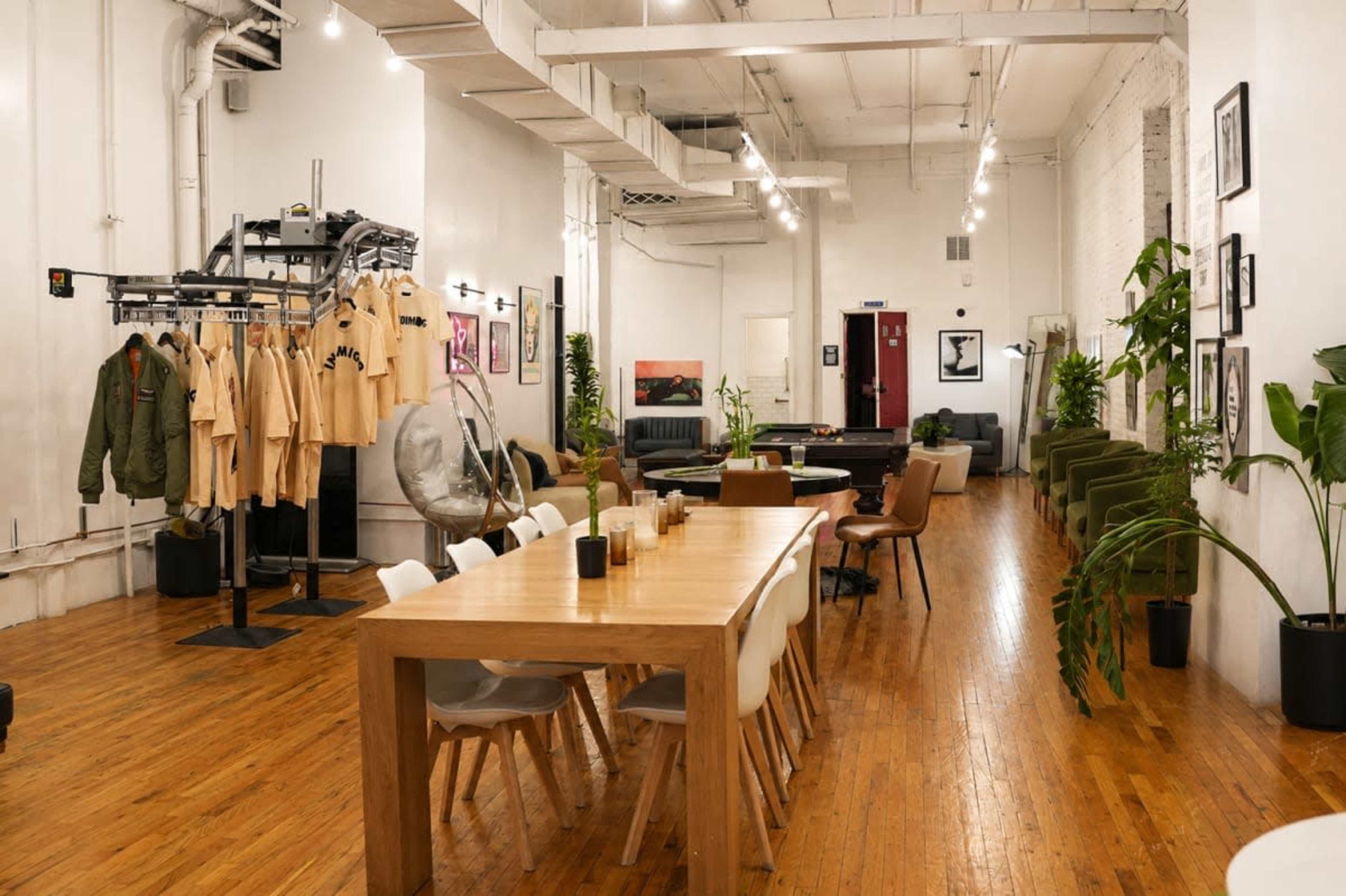 The image shows a spacious, modern interior featuring a wooden dining table, stylish chairs, clothing displays, and greenery throughout the space.