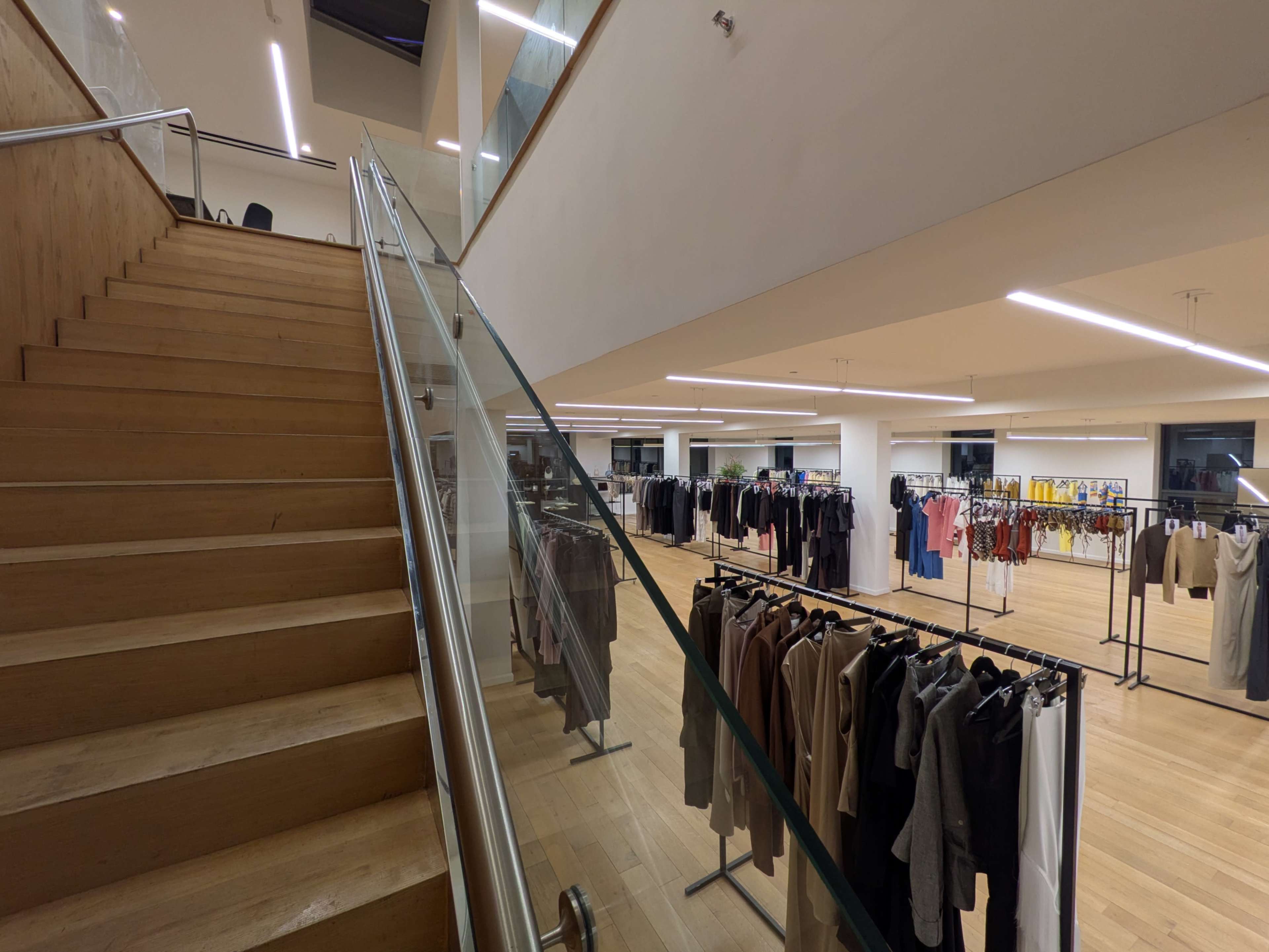 The image shows a modern retail clothing store interior with wooden stairs leading to an upper level and racks of clothing on display.
