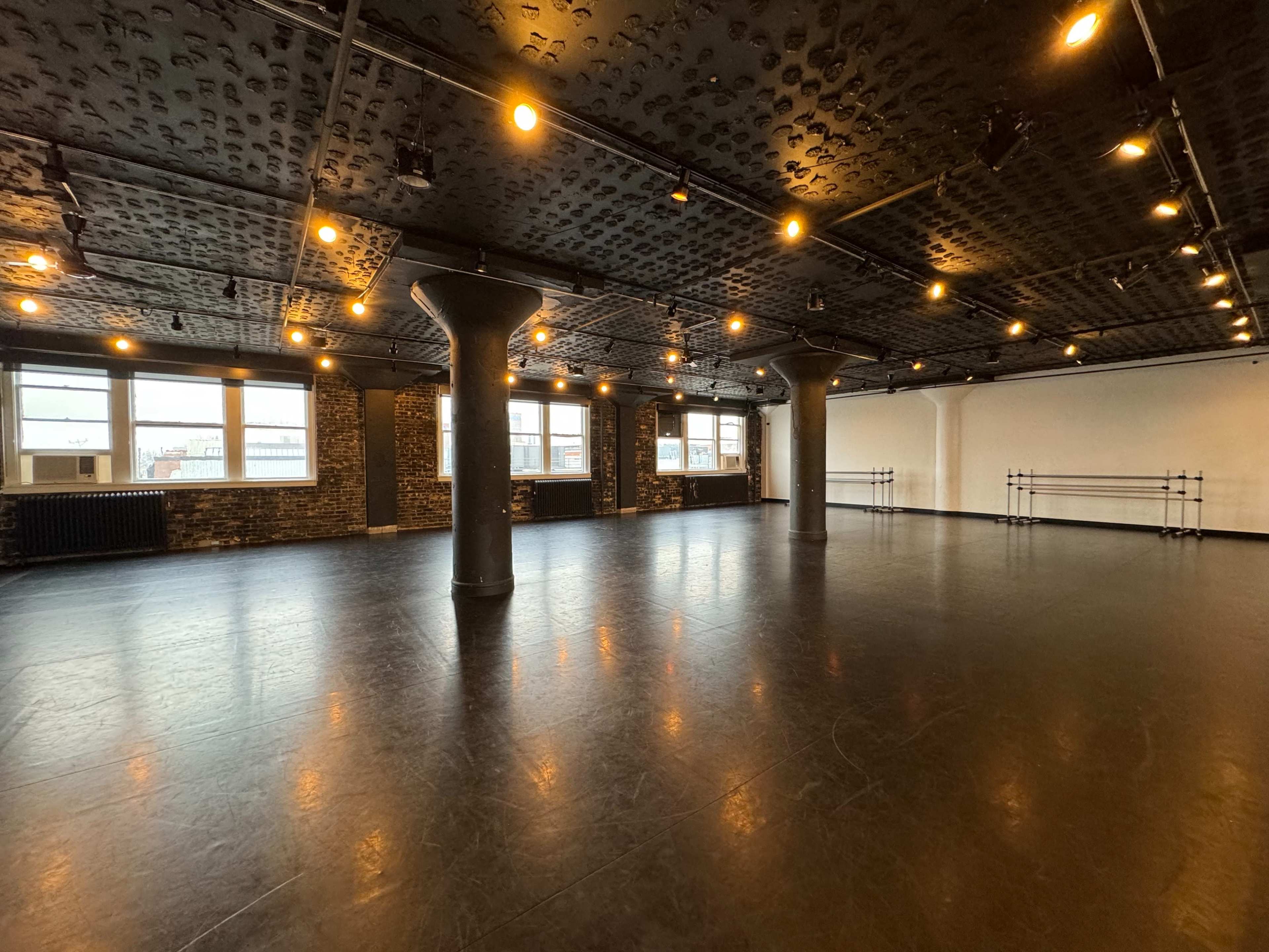 A spacious dance studio with polished black flooring, exposed brick walls, and large windows.