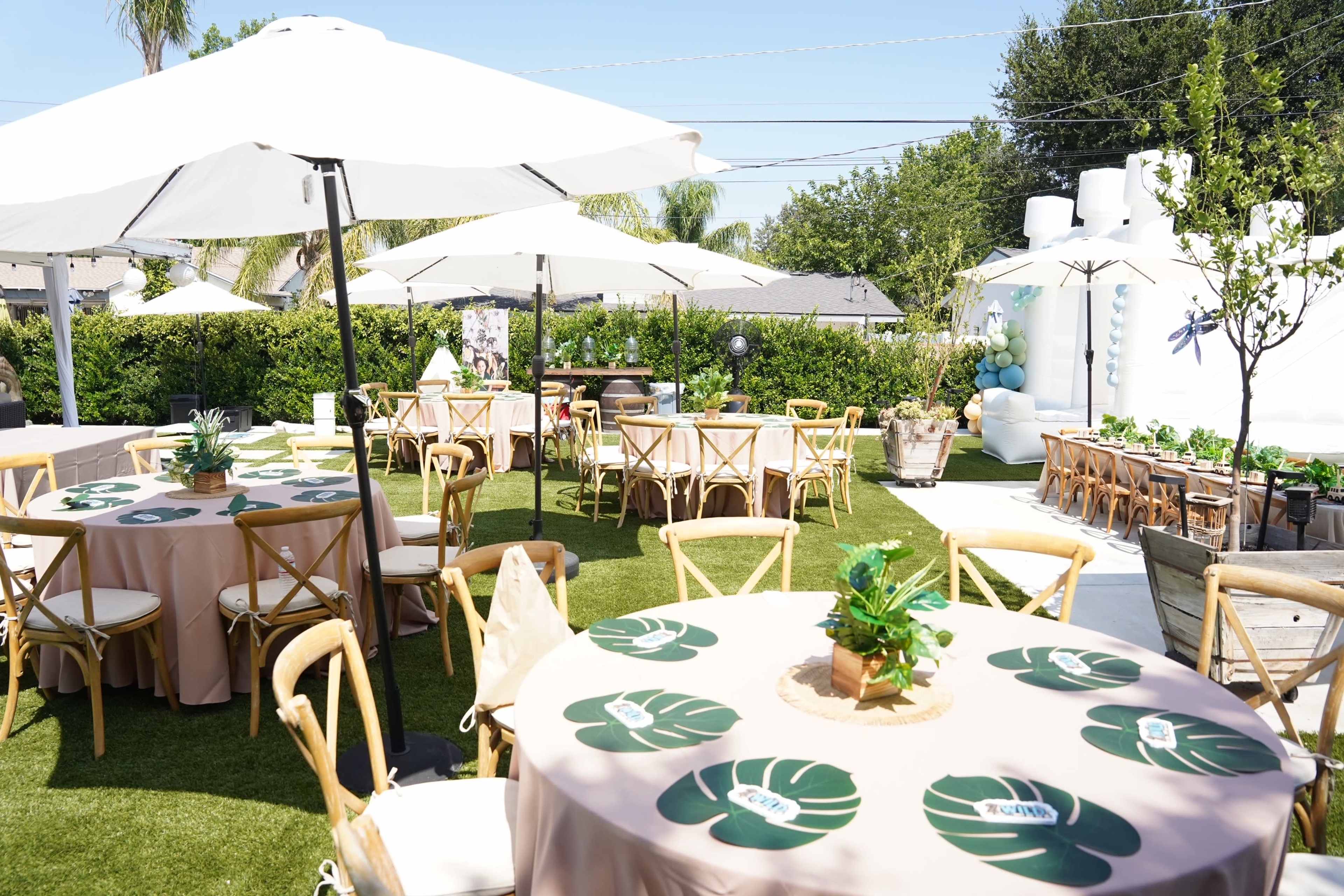 The image shows an outdoor event setup with tables covered in light brown cloth, centerpieces with greenery, and large umbrellas providing shade.