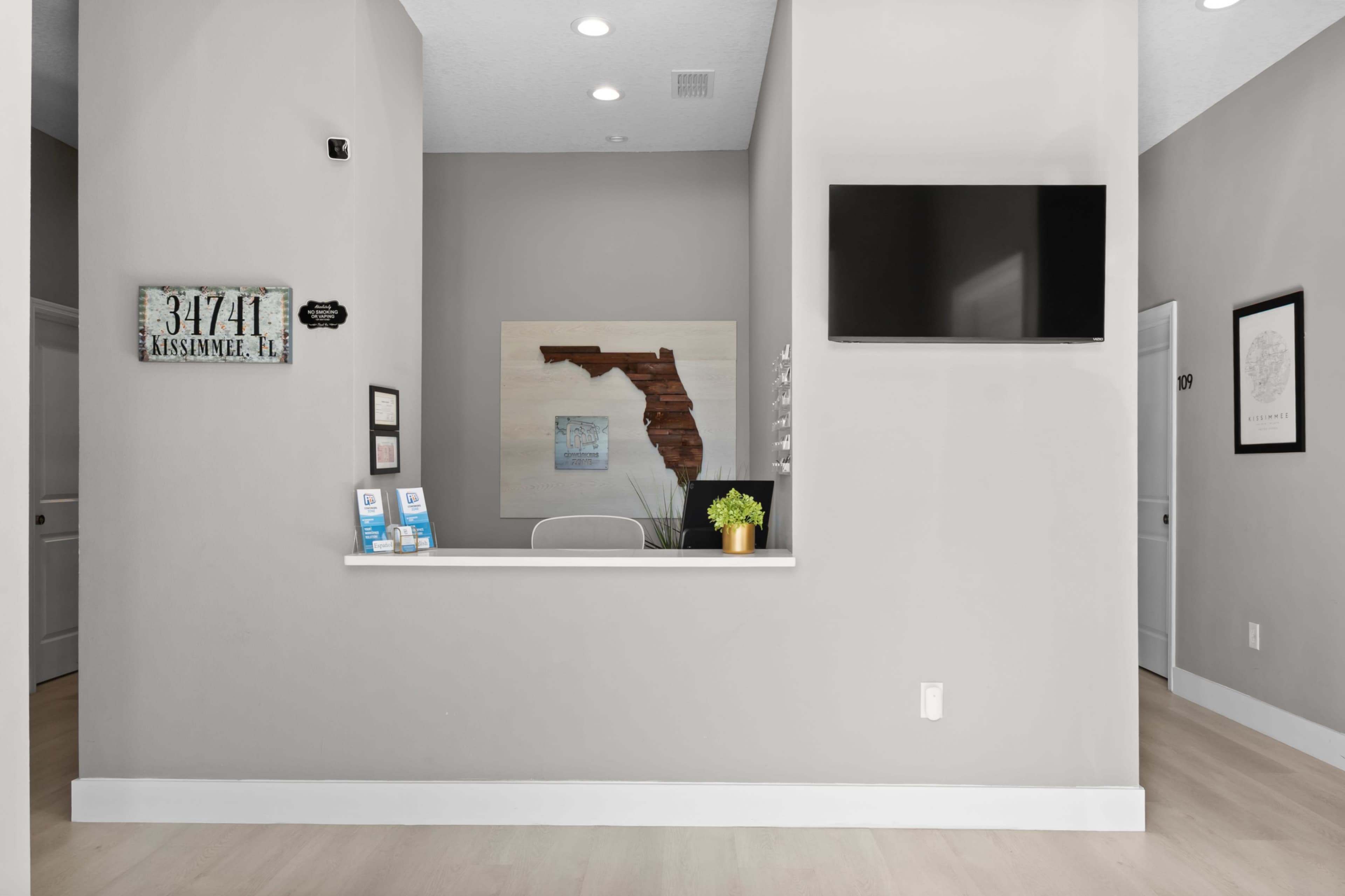 The image shows a reception area featuring a gray wall, a wall-mounted television, a small desk with informational brochures, and a decorative Florida map.