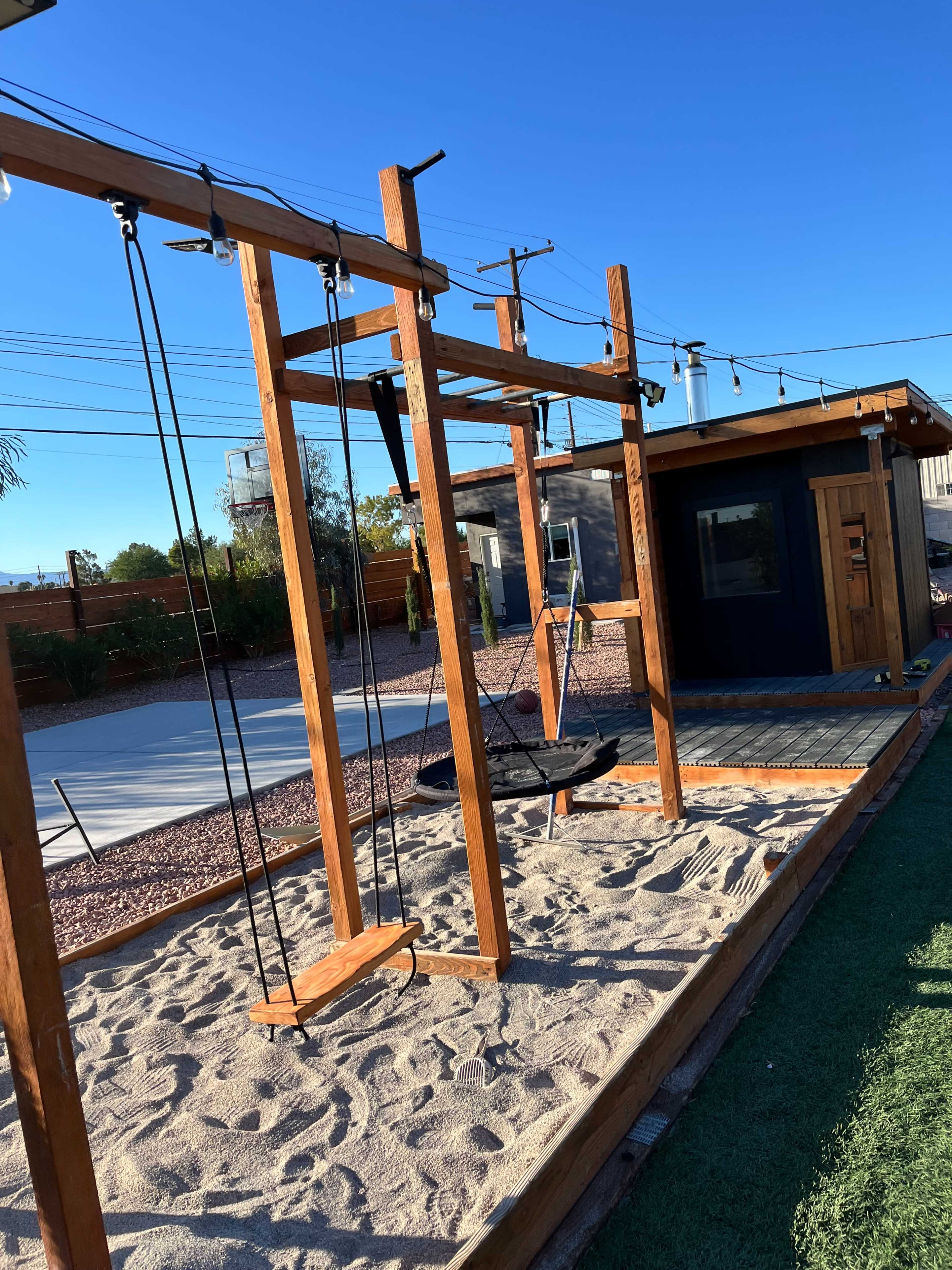 A wooden swing set with a sand base is situated beside a small building in a sunny outdoor area.