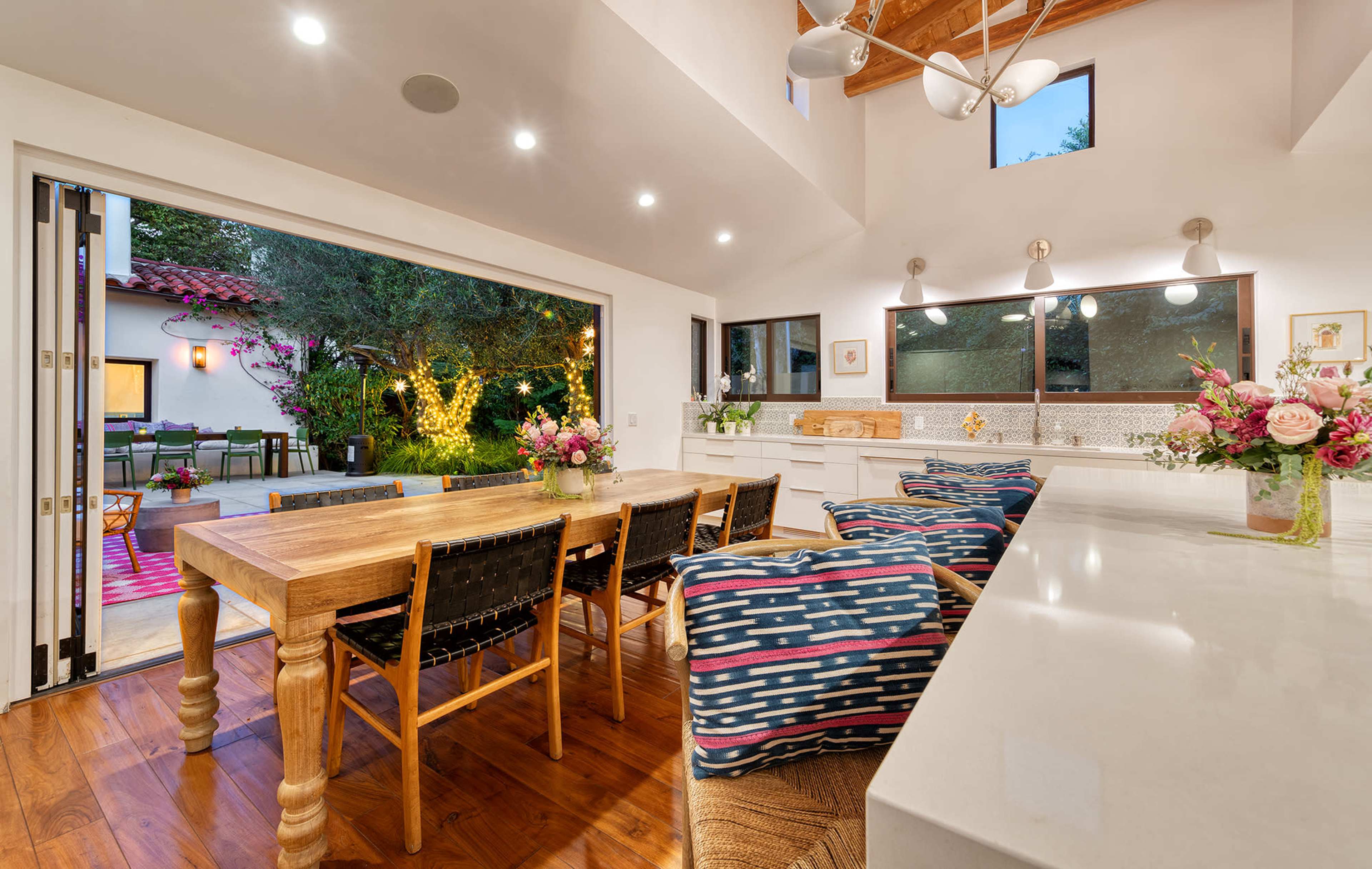 A modern kitchen features a wooden dining table and chairs, large windows opening to an outdoor patio with greenery and decorative lighting.