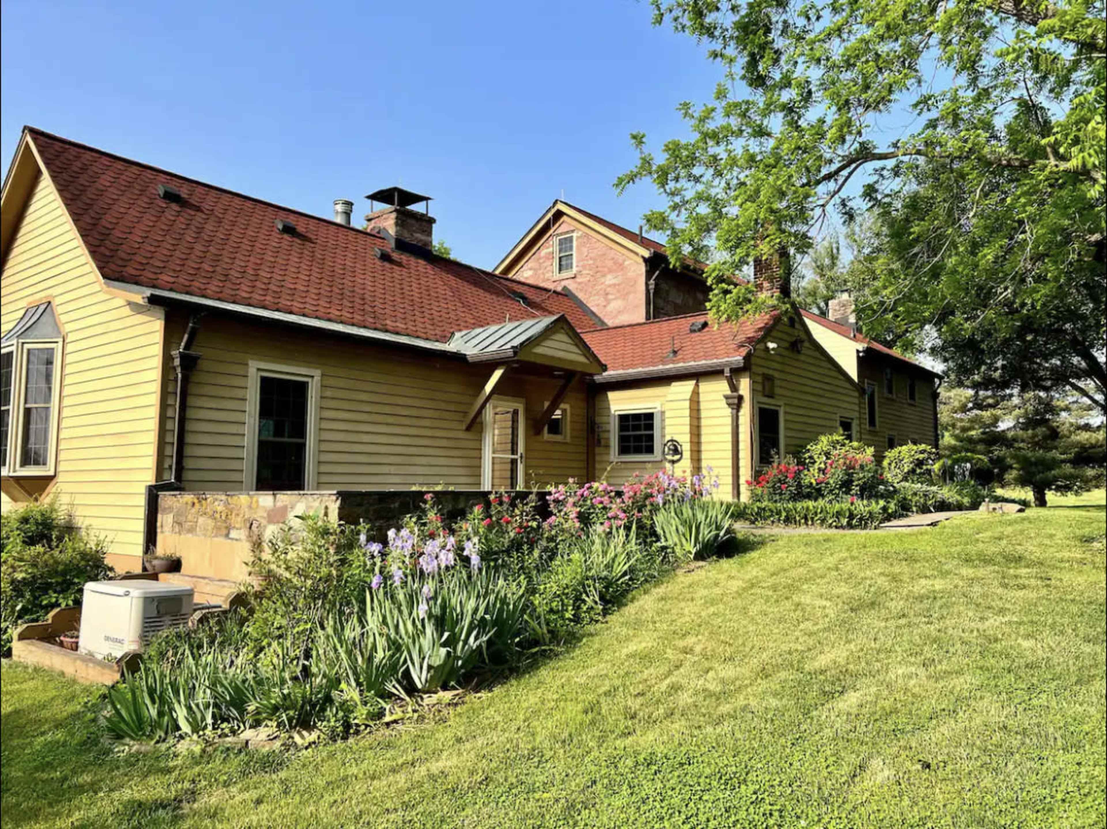 A yellow farmhouse with a red roof is surrounded by green grass and flower beds under a clear blue sky.