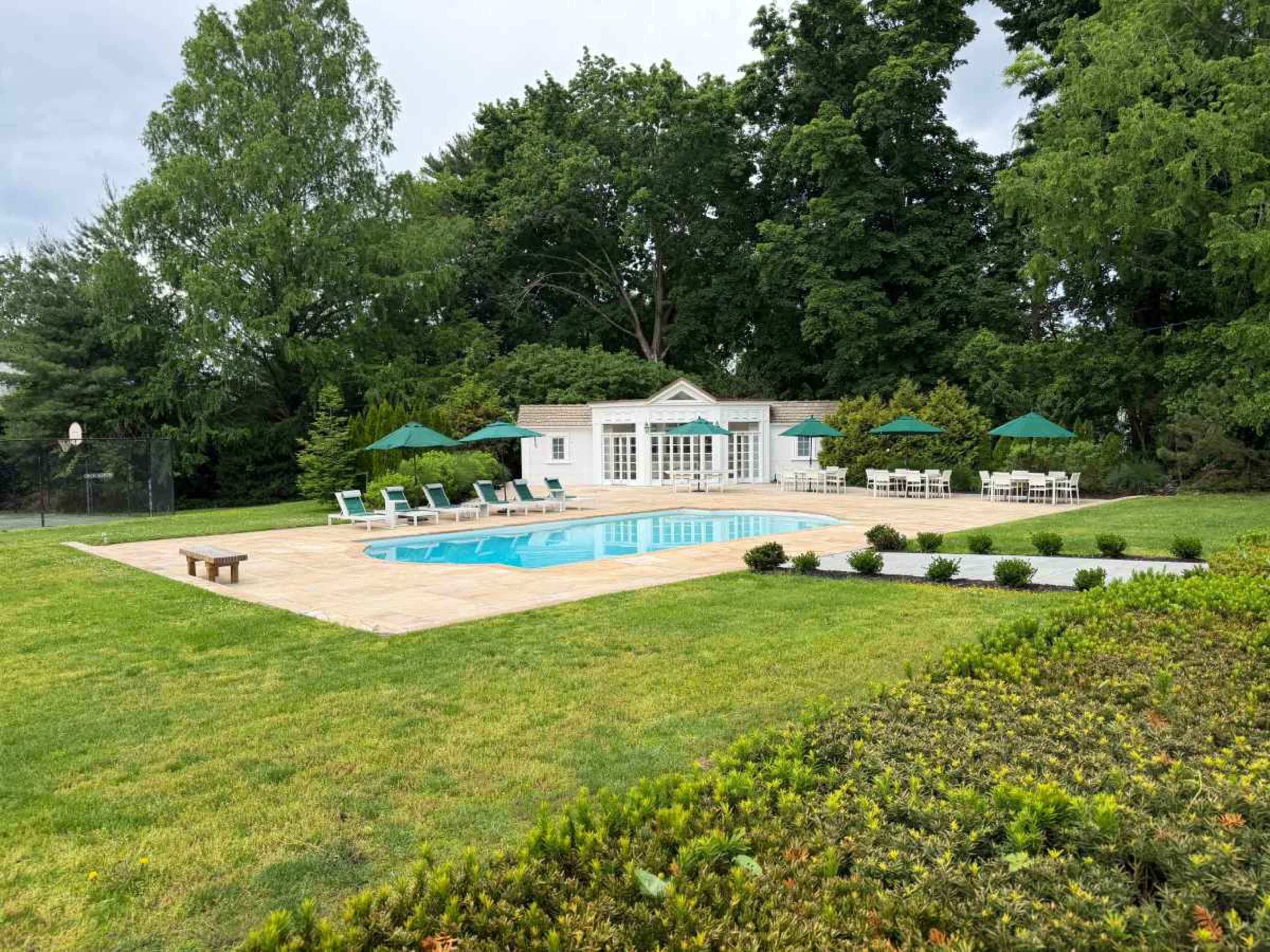 A rectangular swimming pool is surrounded by lounge chairs and green umbrellas in a grassy area beside a white building.