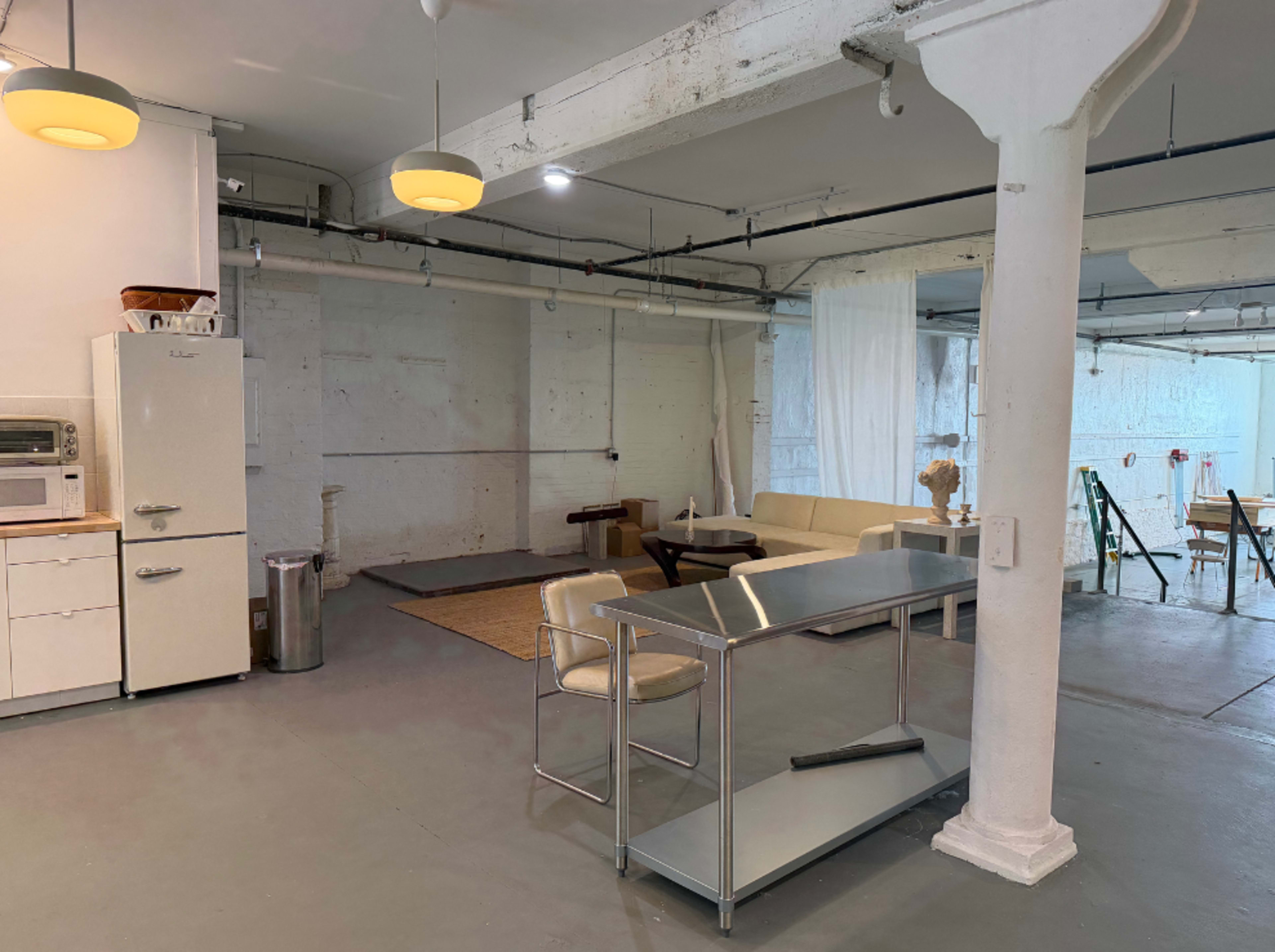 The image depicts an open, industrial-style space featuring a kitchen area with appliances, a table, a sofa, and a distinct separation between the kitchen and living areas.