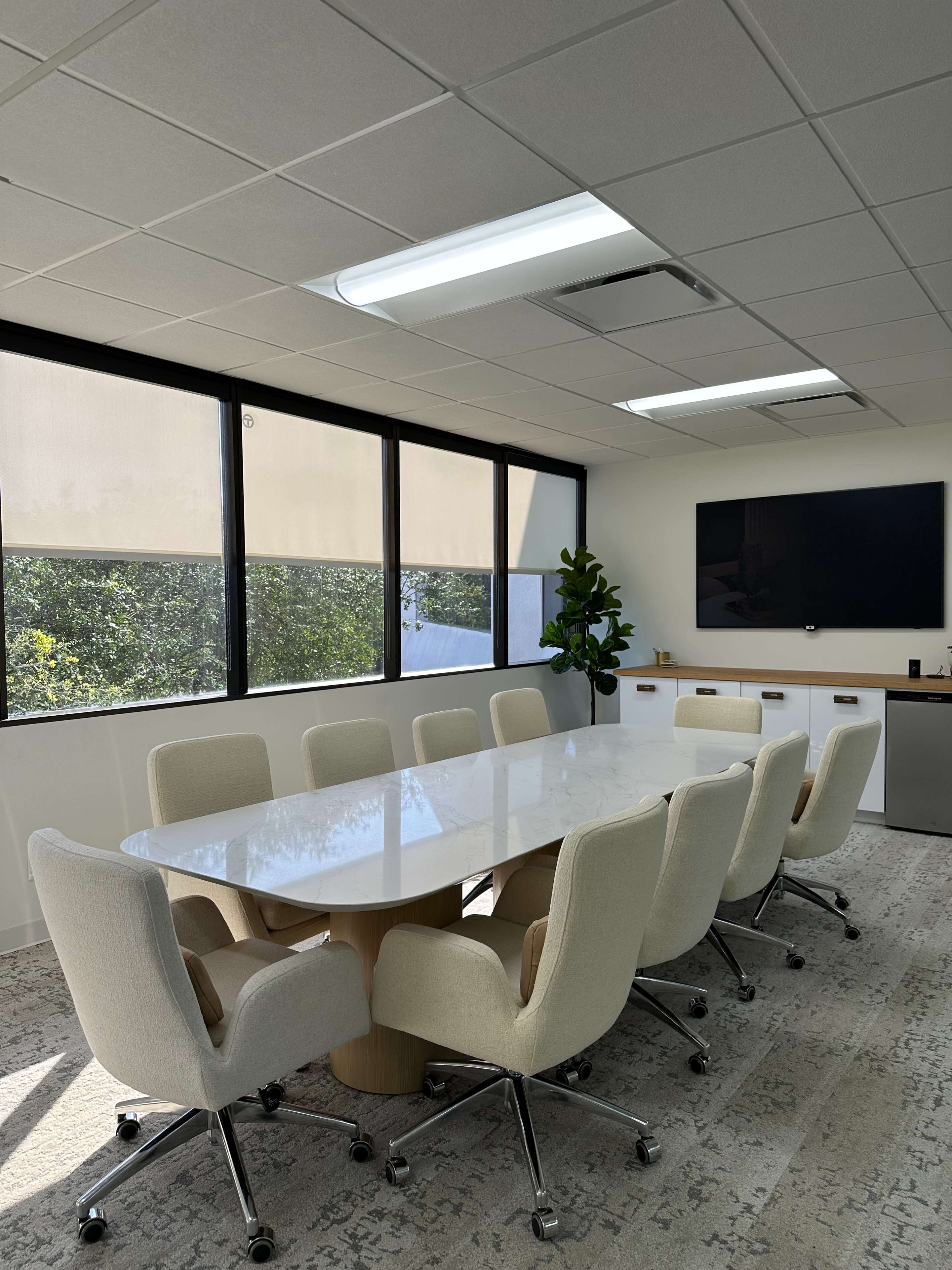 The image shows a modern conference room with a large oval table surrounded by eight chairs, large windows, and a television mounted on the wall.
