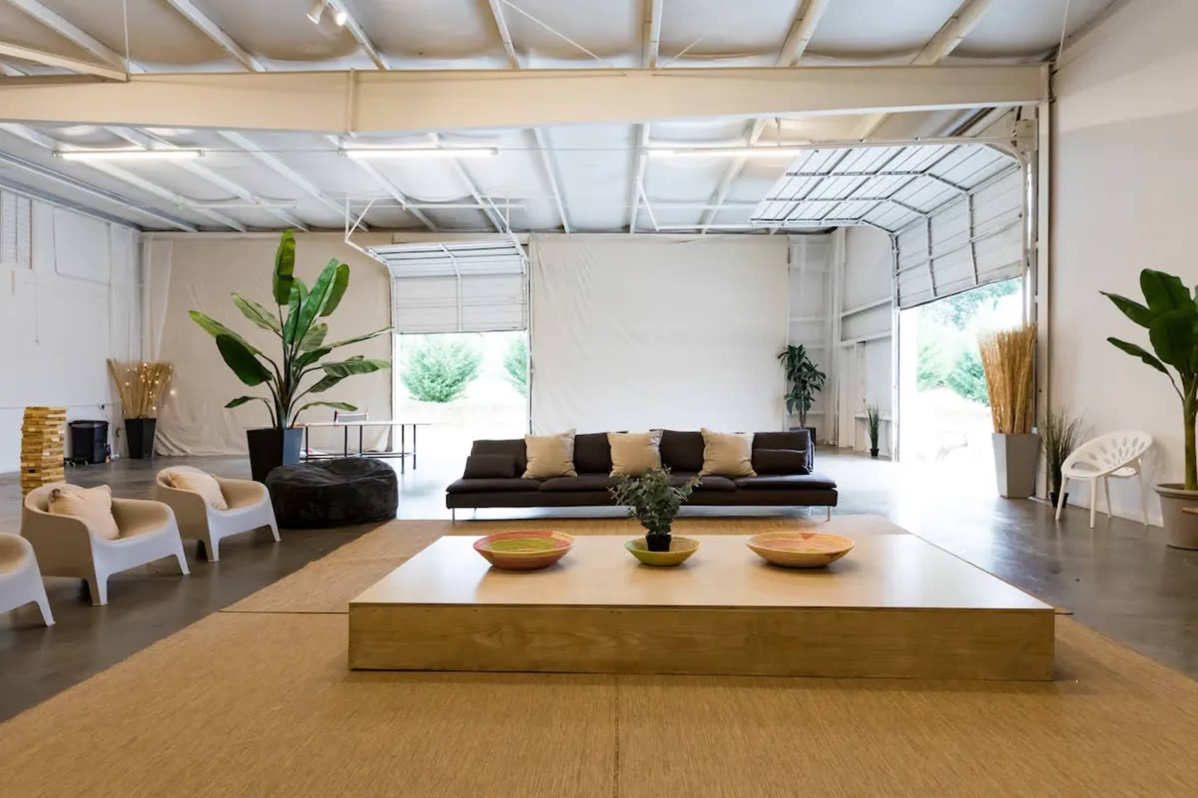The image shows a spacious, modern interior with a large sofa, a wooden coffee table, and plants arranged around a light-colored rug.