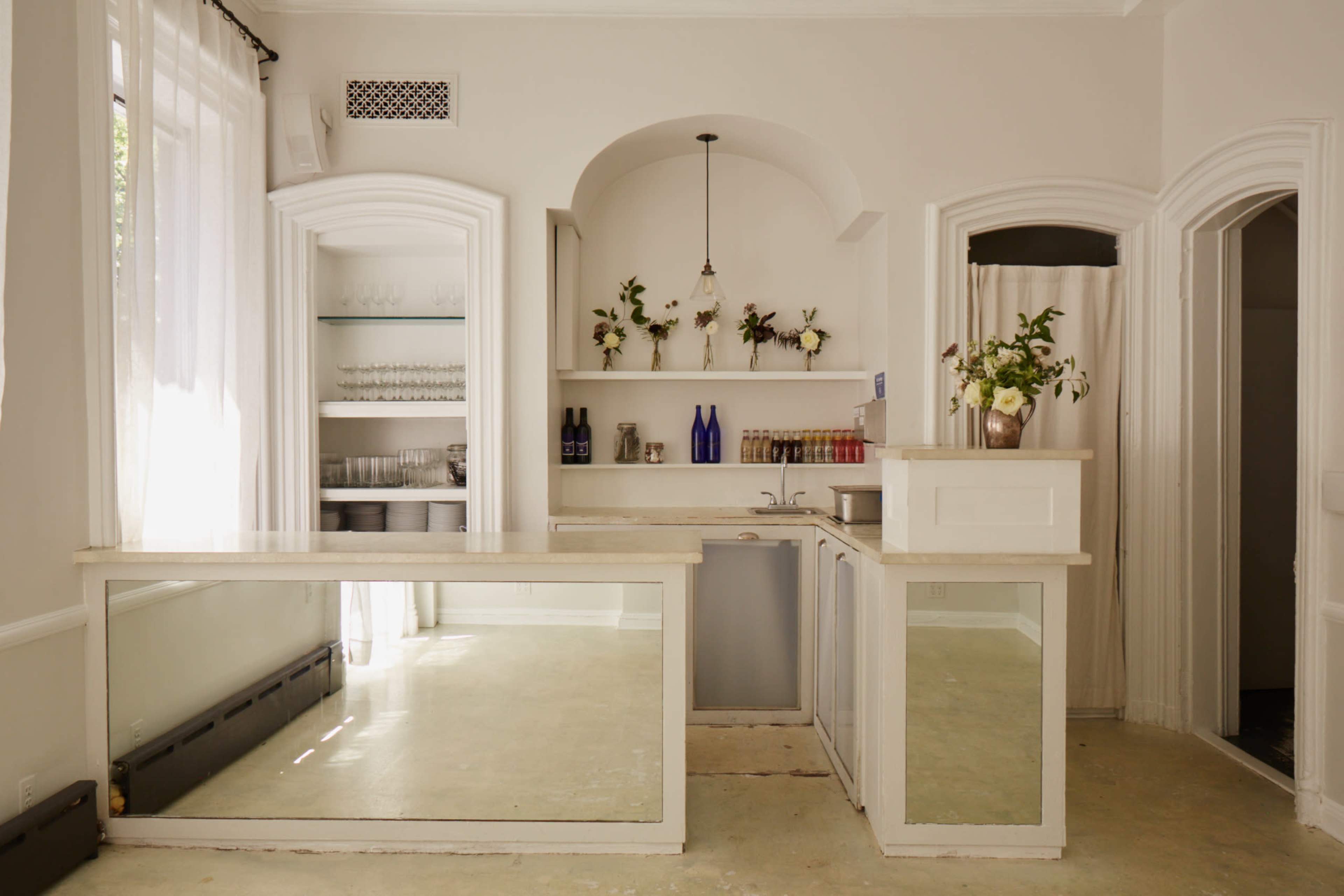 The image features a minimalist room with a clean bar area, wood countertops, and shelves displaying glassware and decorative items.