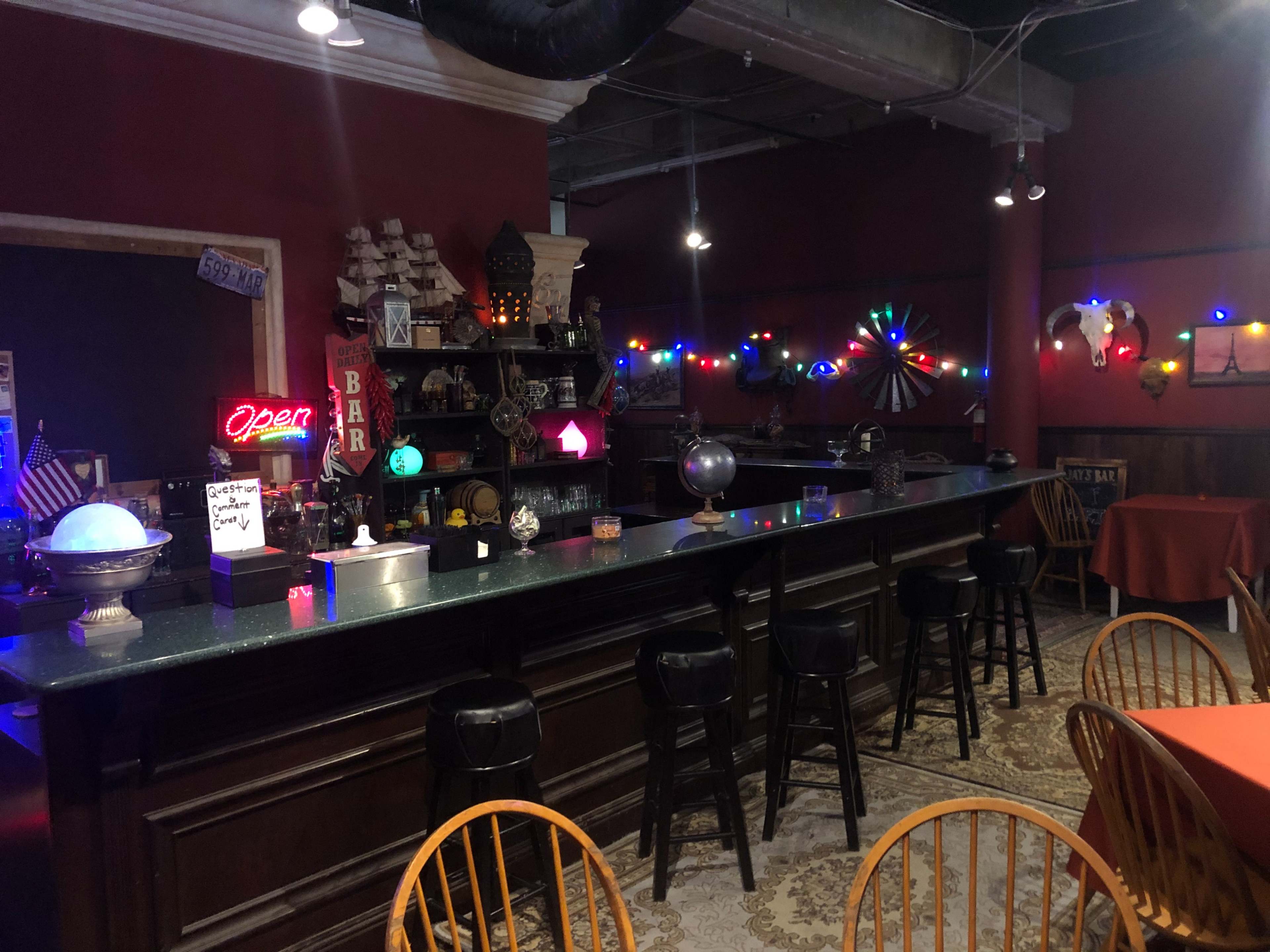 The interior of a bar with a long, dark countertop, several high stools, decorative lights, and various ornaments on shelves against a red wall.
