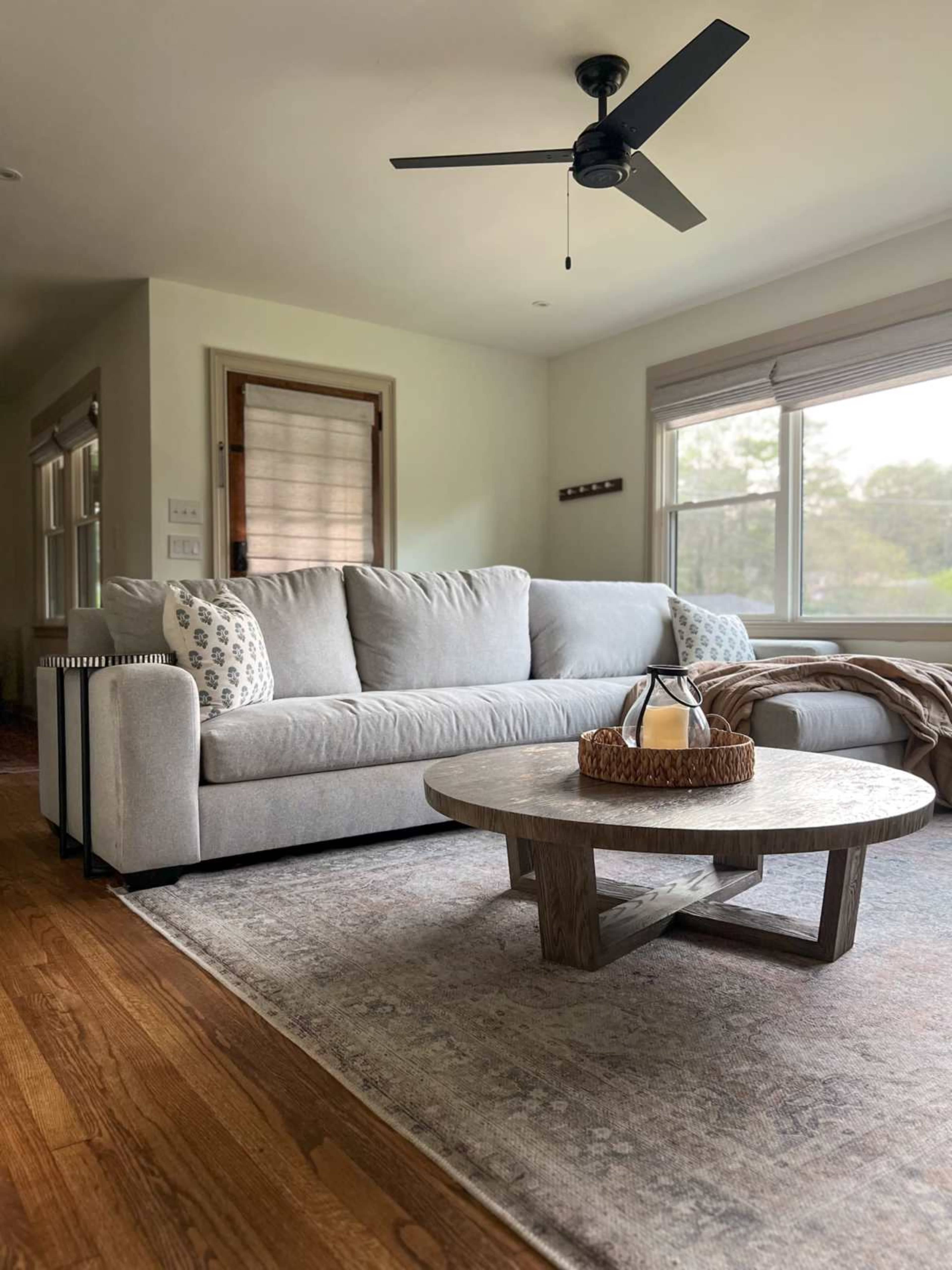 A light-colored sofa with decorative pillows is positioned in a living room, accompanied by a round wooden coffee table and a ceiling fan.