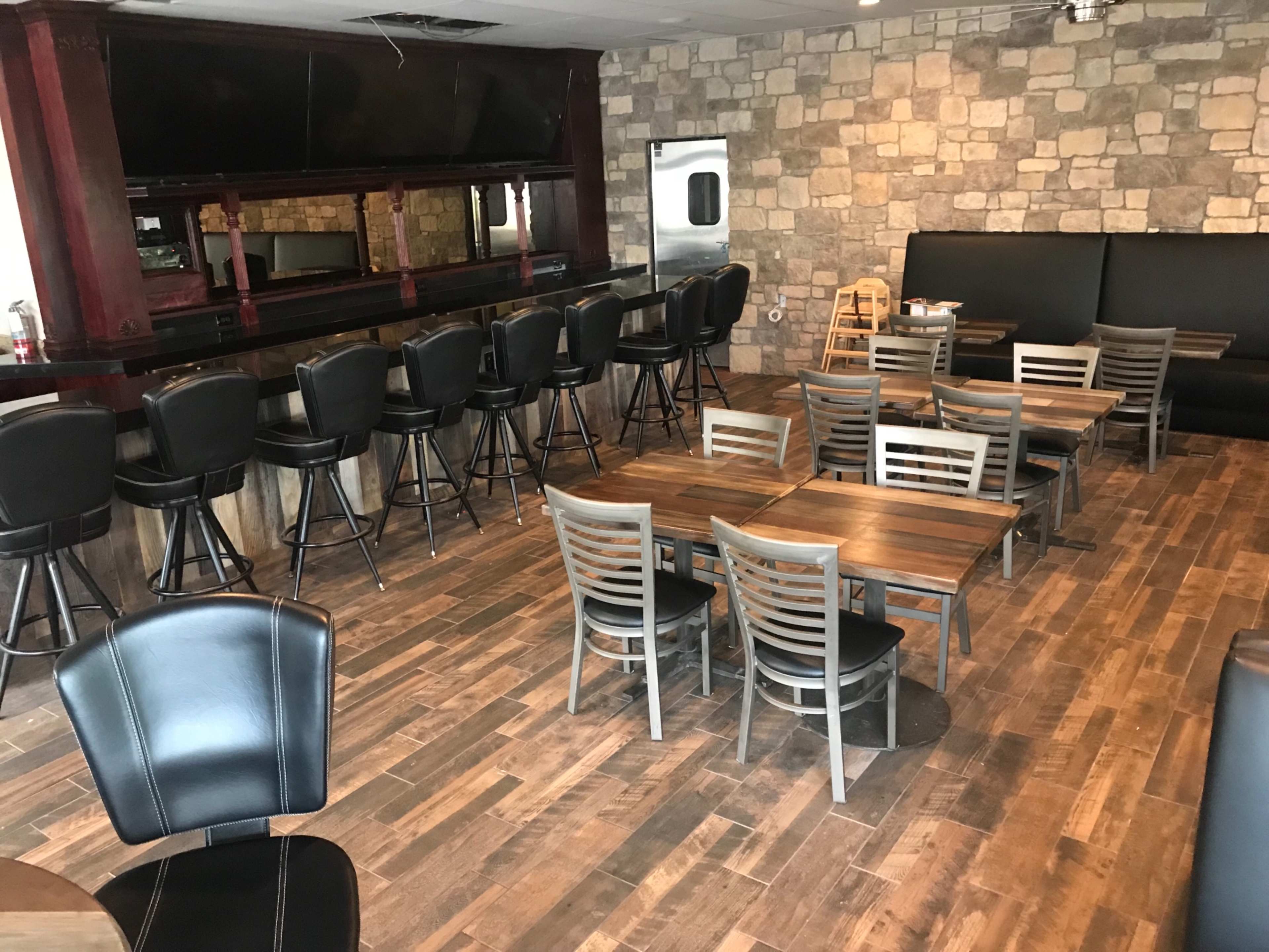 A restaurant interior featuring a bar with high stools, several tables with chairs, and a stone-patterned wall.