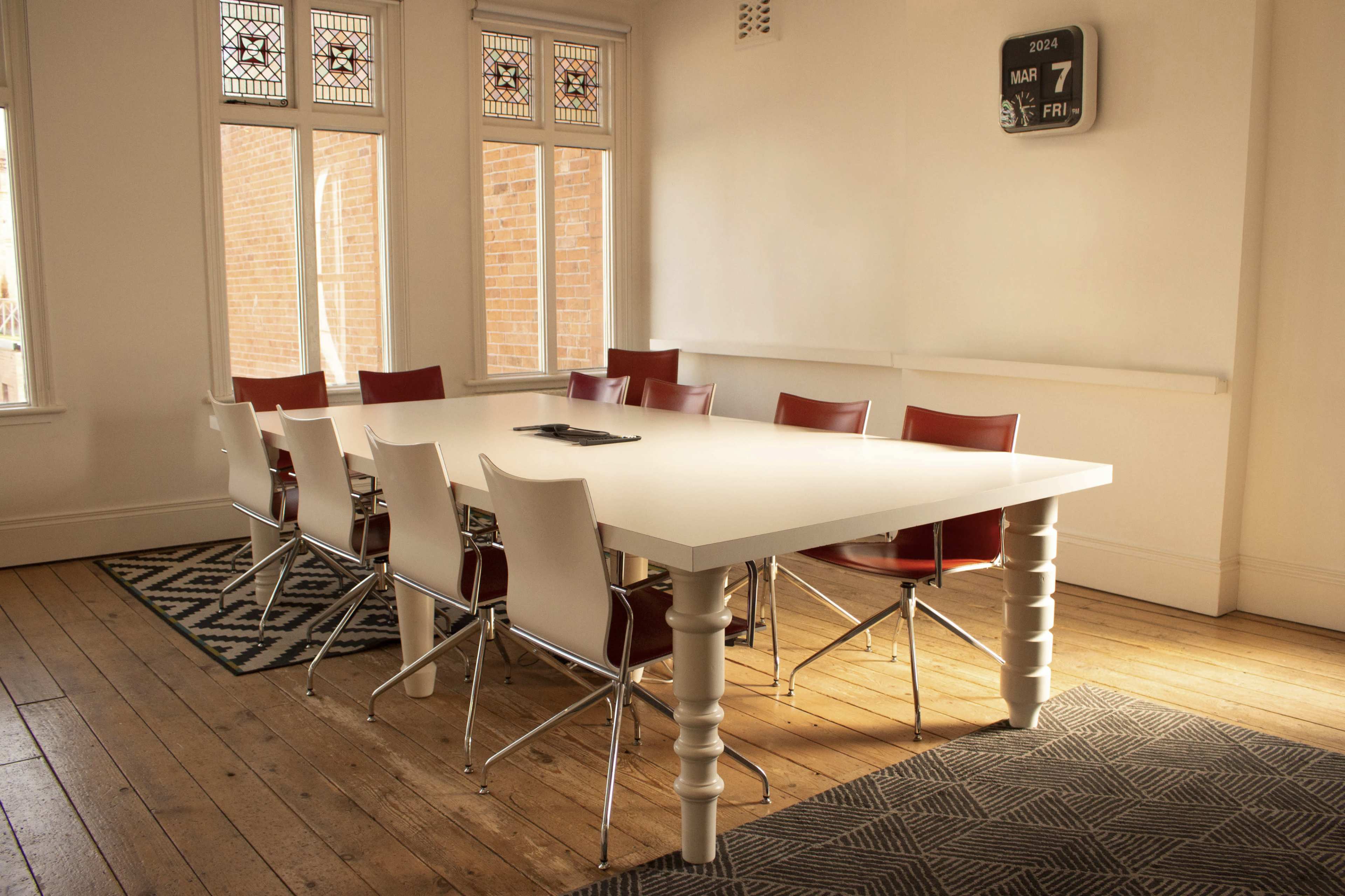 A large white table with several red and white chairs is positioned in a well-lit meeting room with wooden floors and large windows.