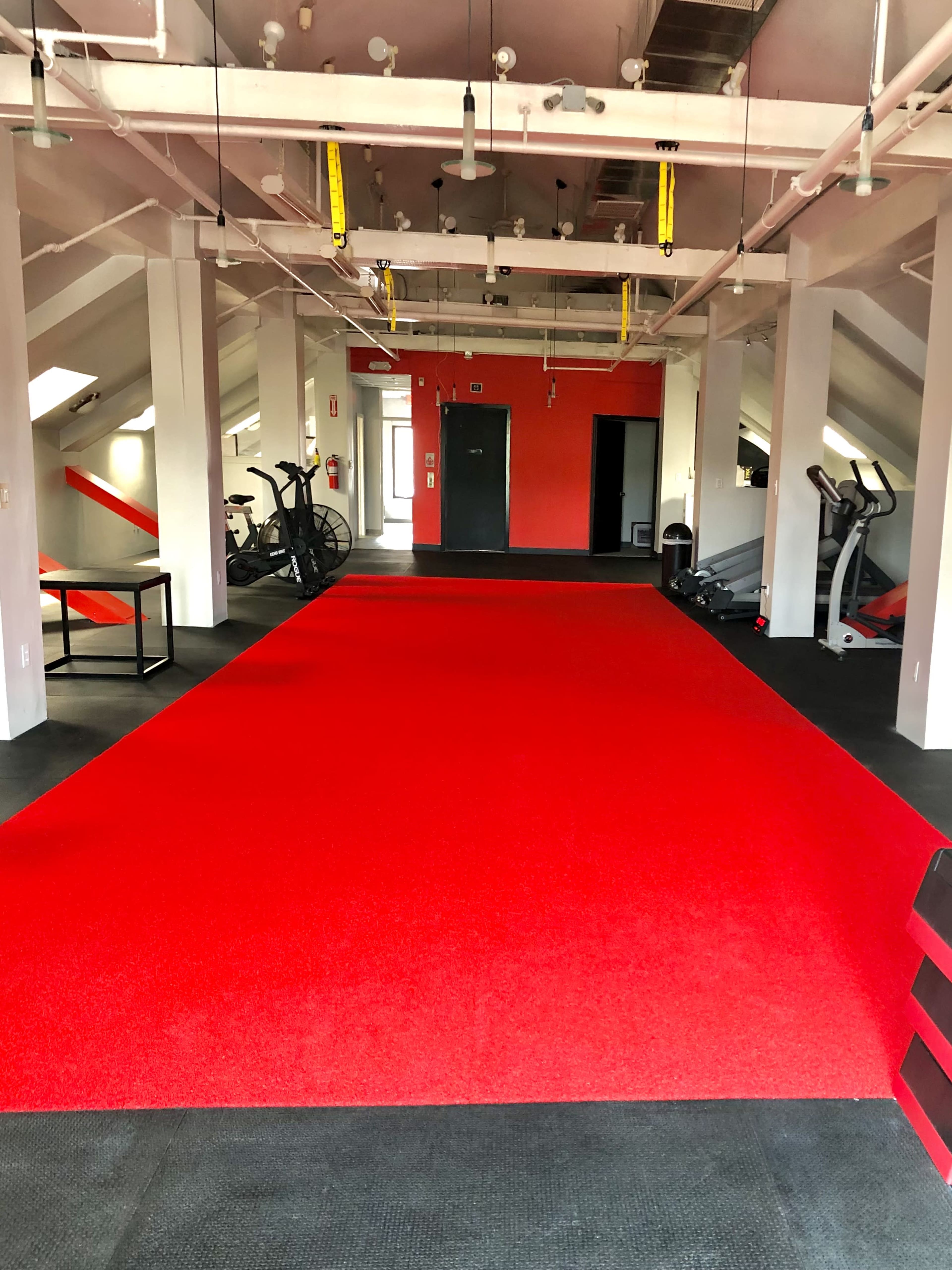 A gym interior with red carpet flooring, exercise equipment, and white pillars in a well-lit space.