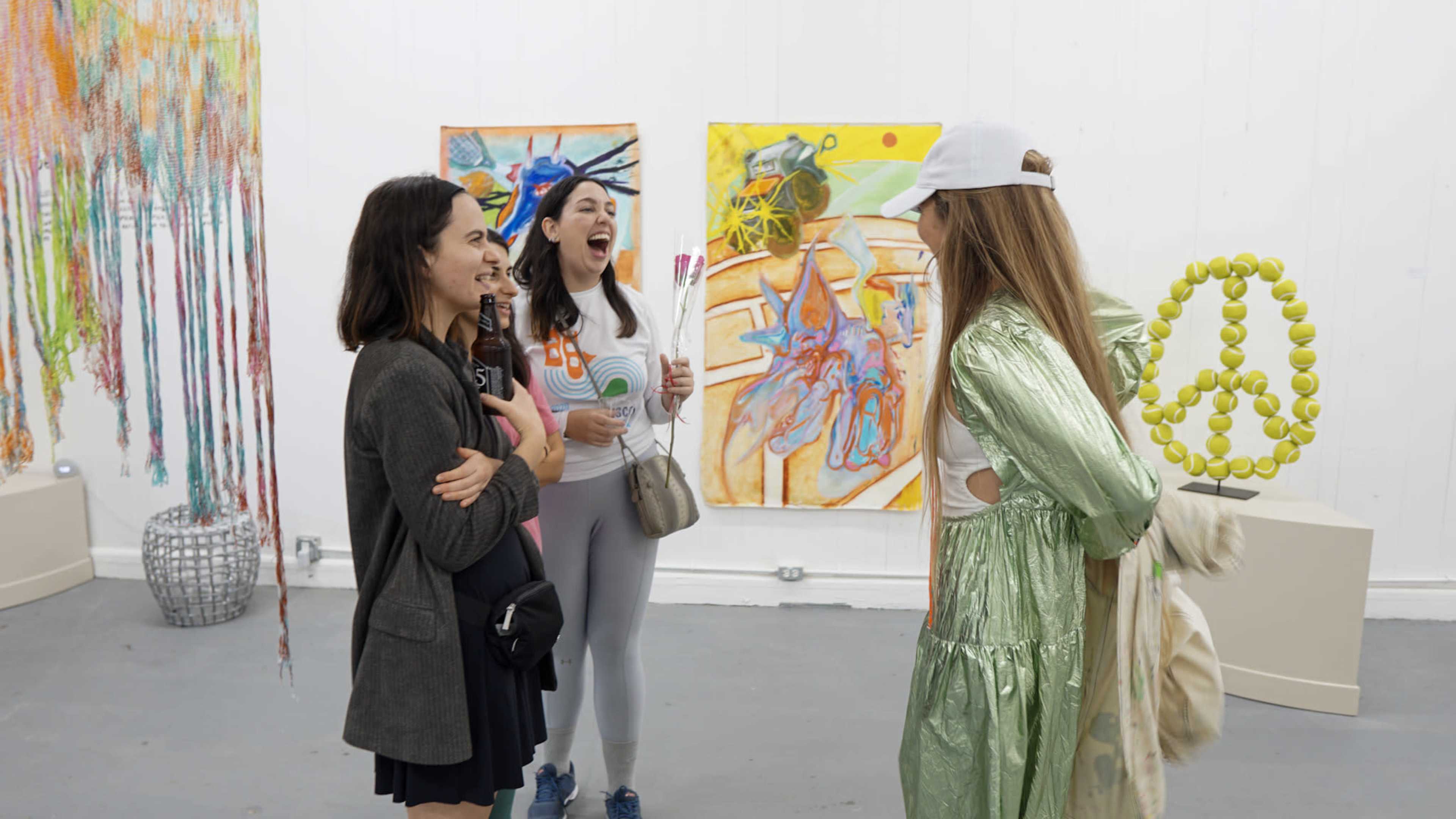 A group of four women engage in conversation in an art gallery filled with colorful abstract paintings.