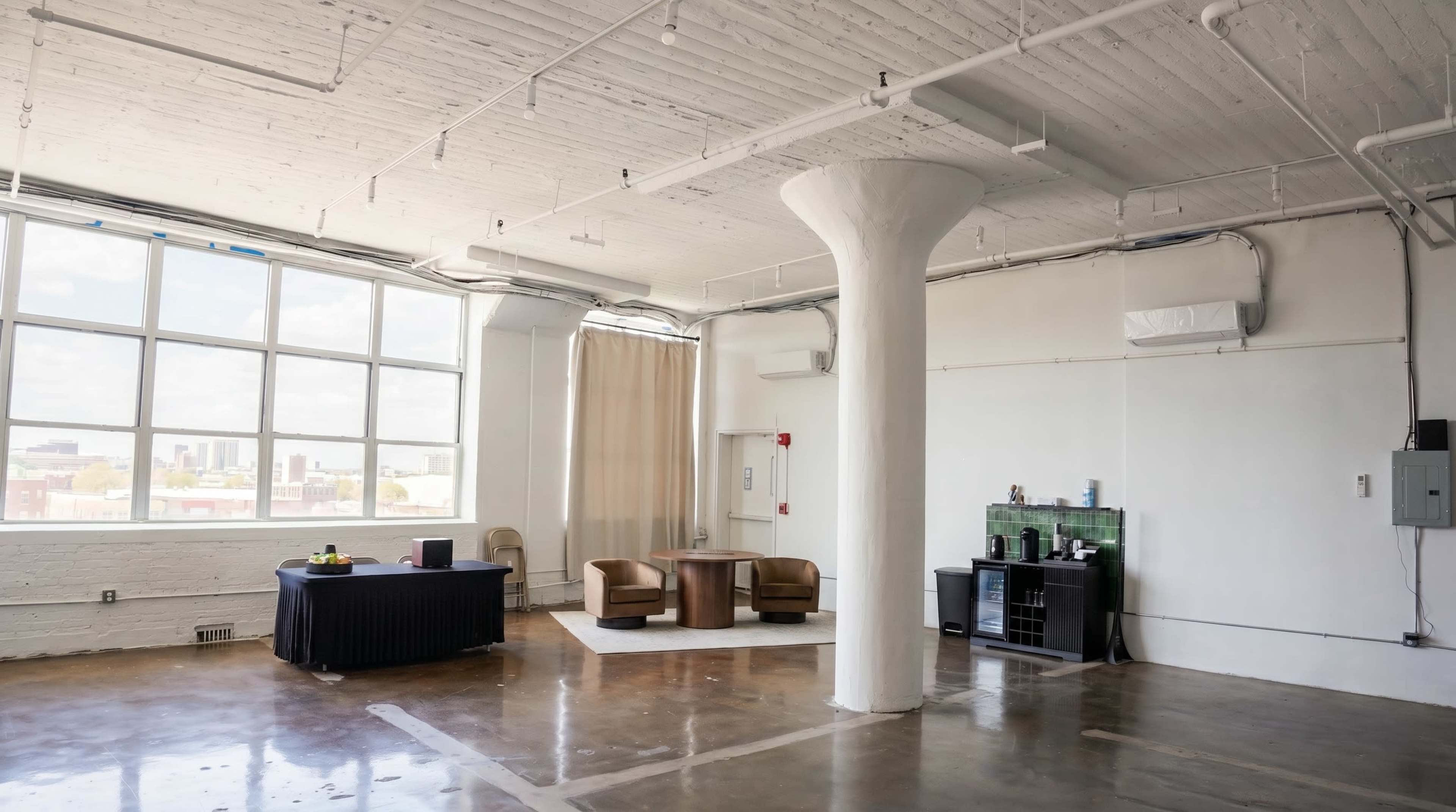 The image shows a spacious, airy room with large windows, featuring a seating area with two chairs and a table, alongside a small kitchenette and a column in the center.