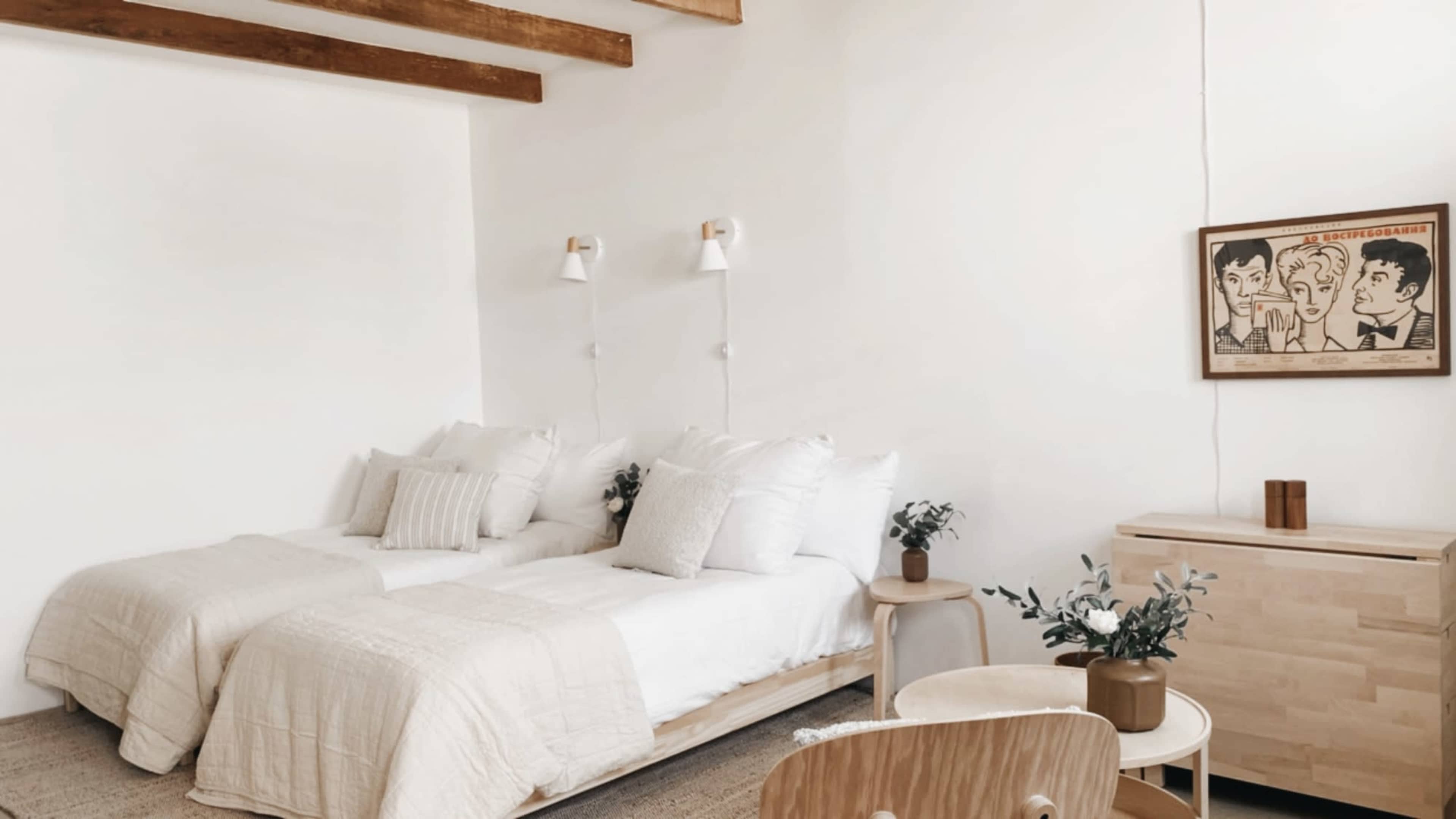 The image shows a minimalist bedroom featuring two twin beds with white bedding, a small wooden nightstand, and a wall-mounted lamp.