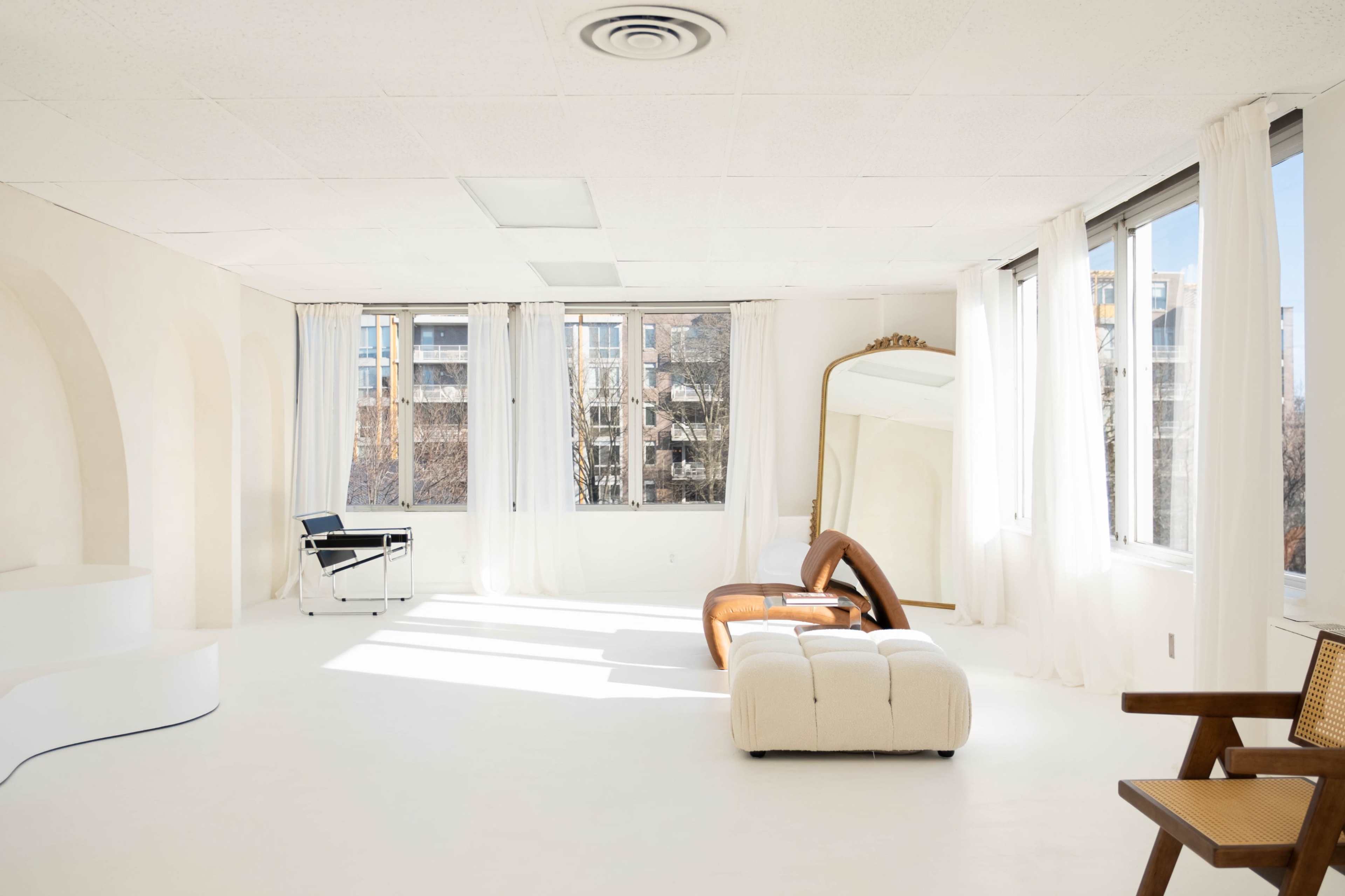 A bright, minimalist room features large windows, white walls, and modern furniture, including a curved sofa and a full-length mirror.