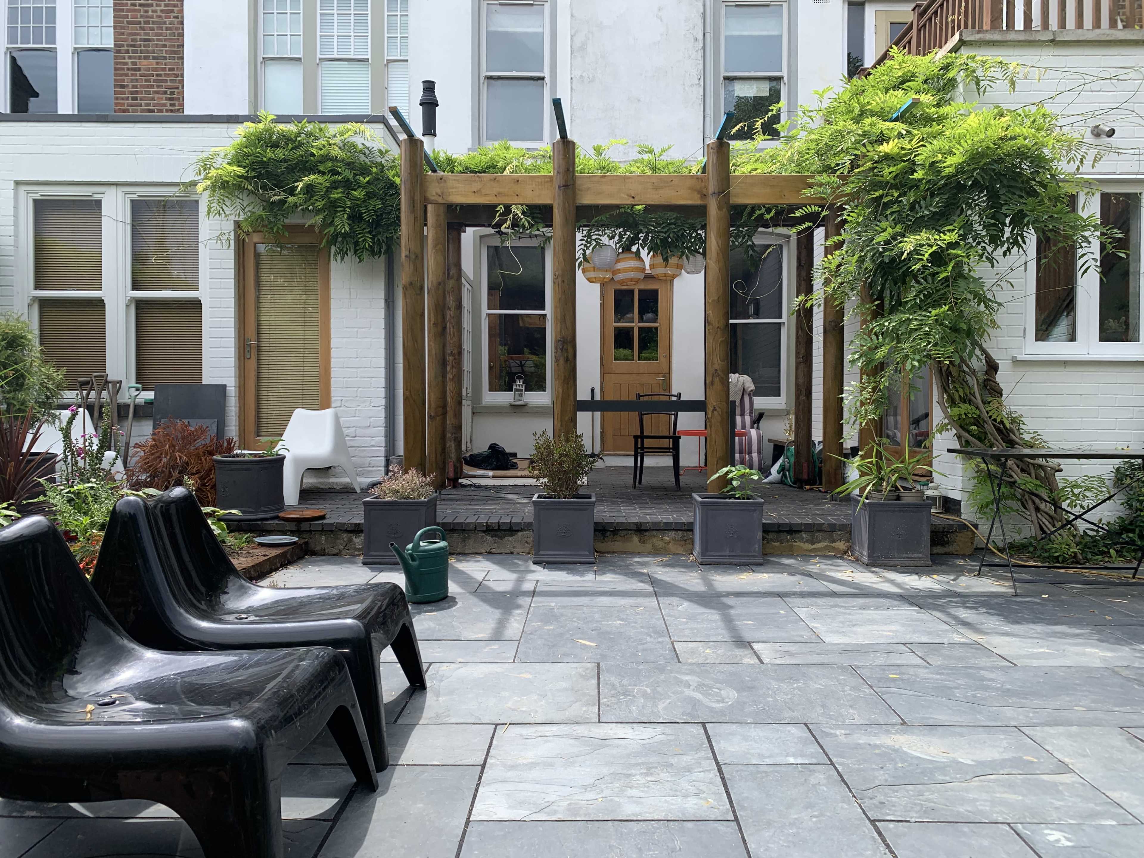 A patio features black chairs, stone flooring, planters, and a wooden pergola covered with greenery.