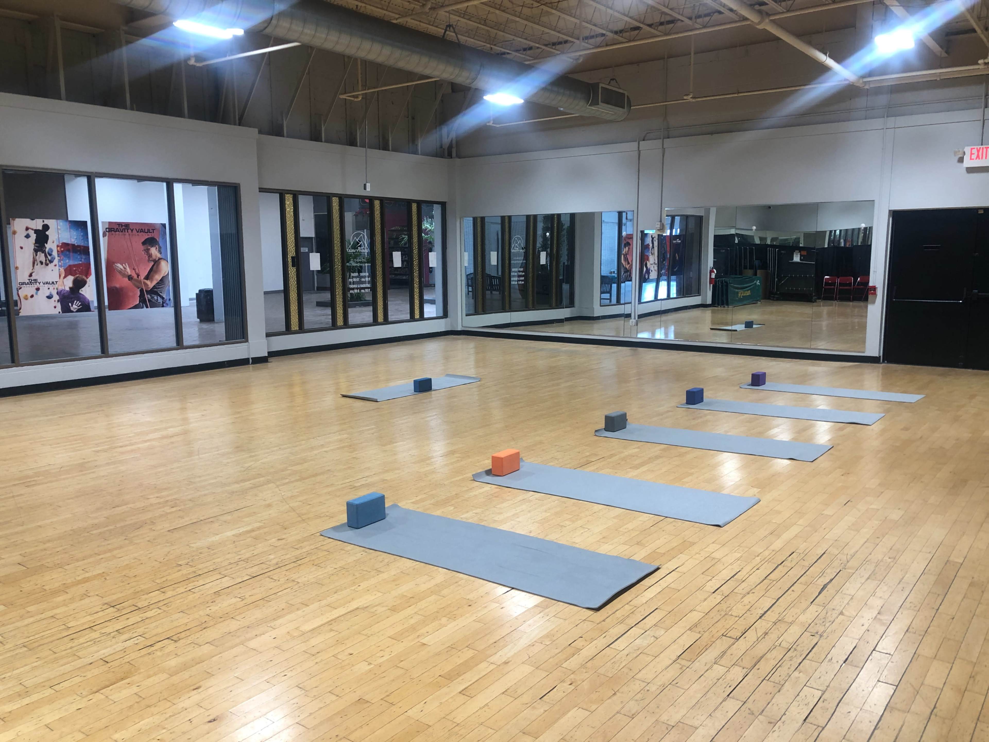 A spacious yoga studio features mats arranged on a polished wooden floor, with blocks placed in front of each mat and mirrors lining the walls.