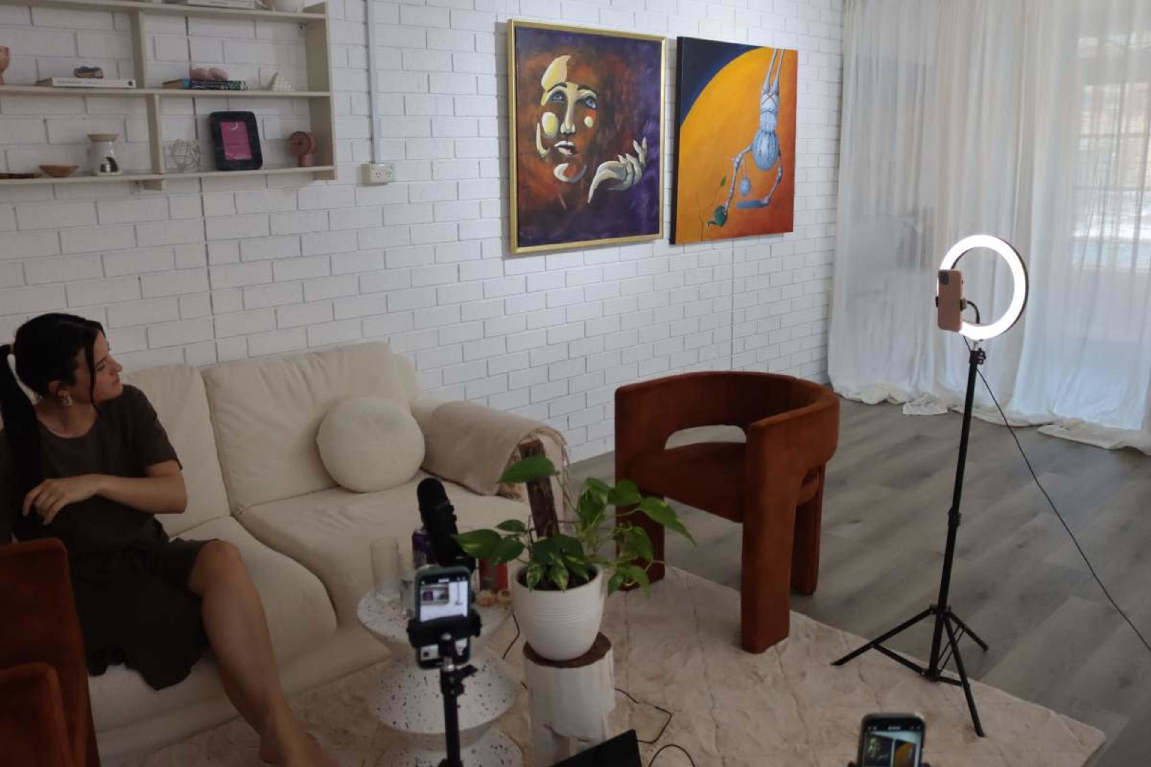 A woman sits on a white sofa in a well-lit room with two paintings on the wall and a ring light on a tripod beside her.