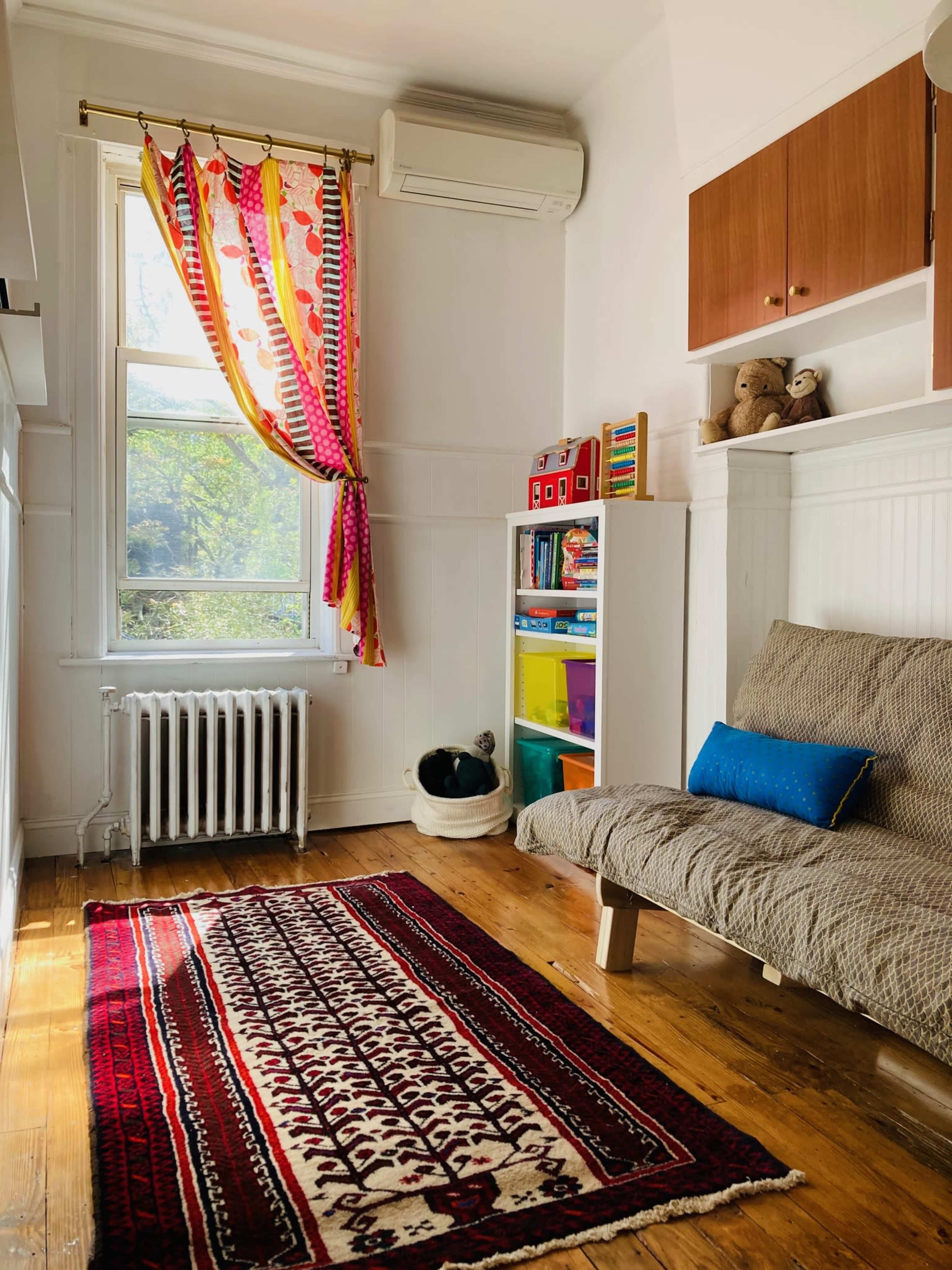 A colorful room features a futon, shelves of toys, a patterned rug, and bright curtains hanging by a window.