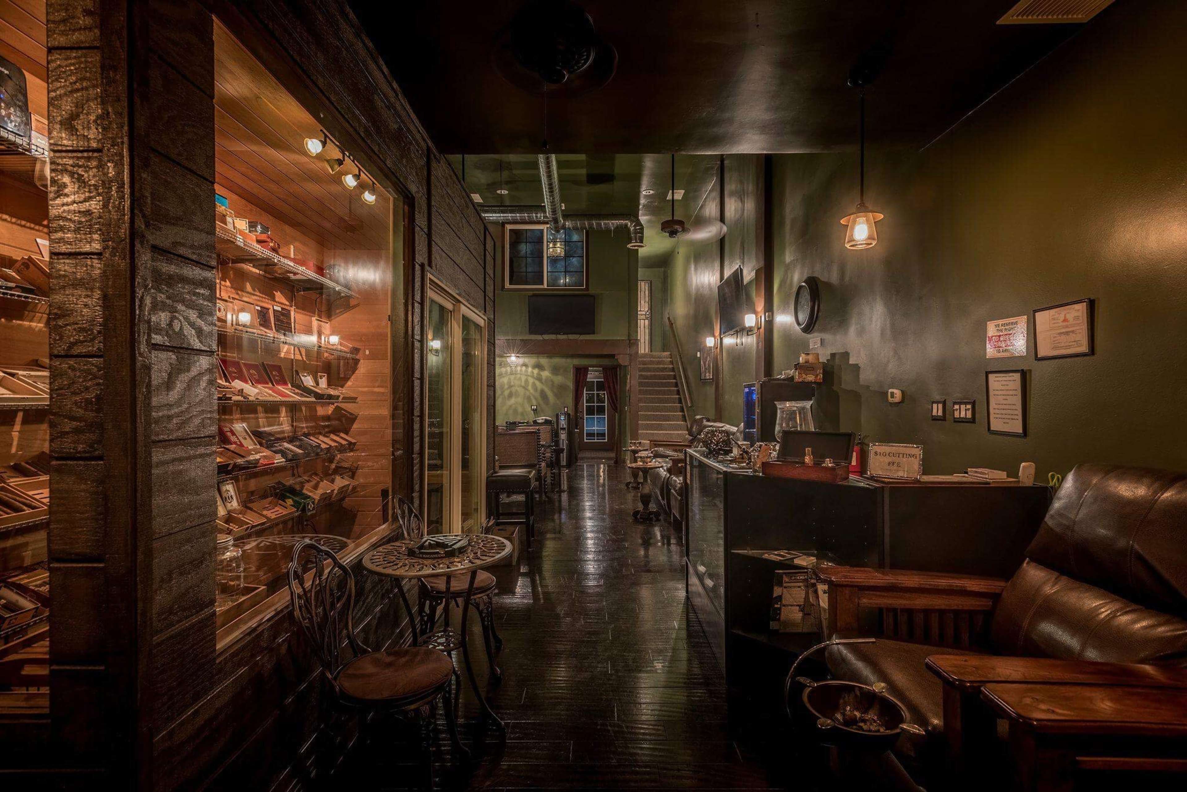 A dimly lit lounge with a cigar display on the left, comfortable seating, and a staircase at the end of a long, narrow hallway.