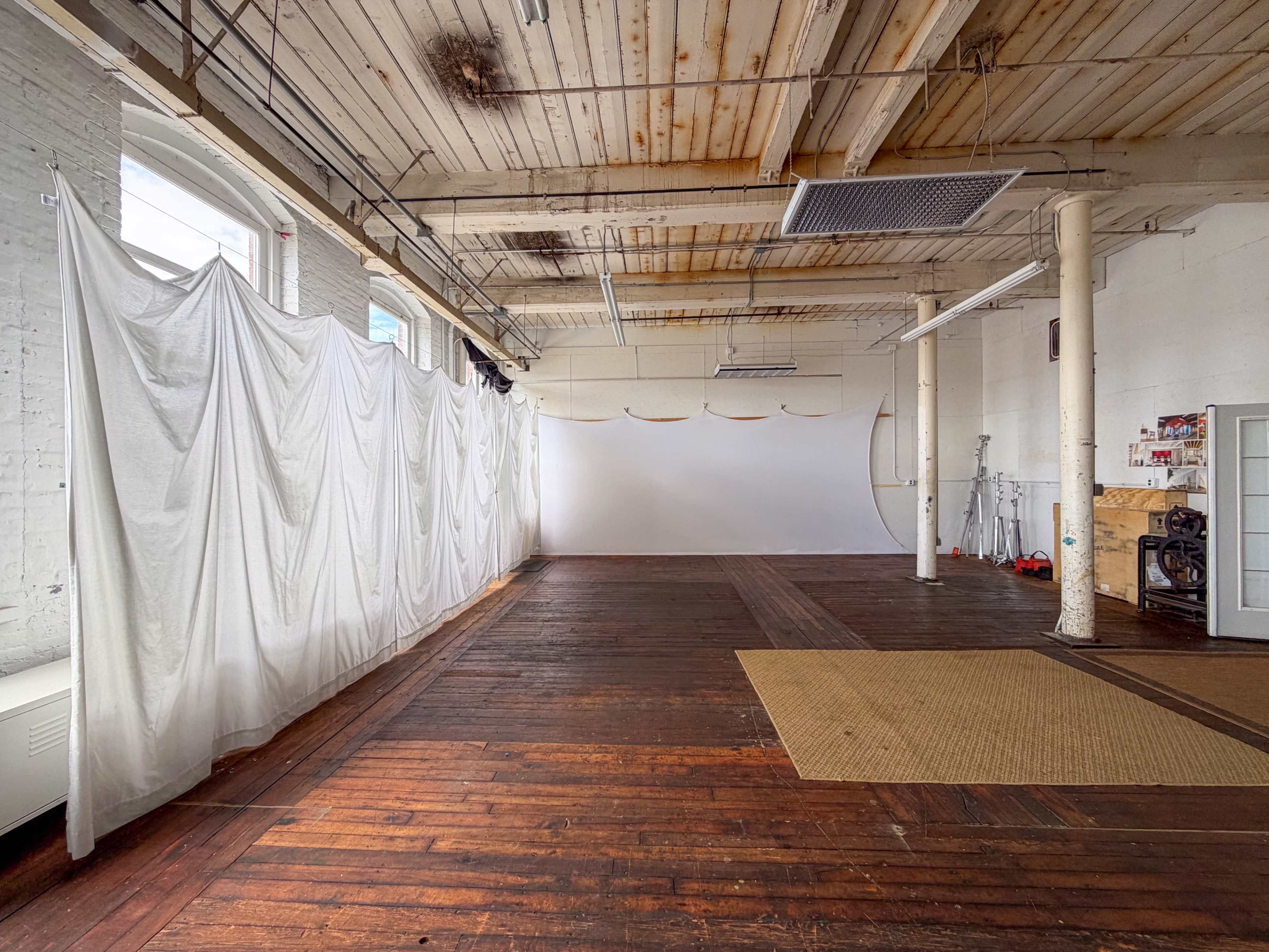 The image shows an empty studio space with wooden floors, white walls, and a fabric divider draped across part of the room.