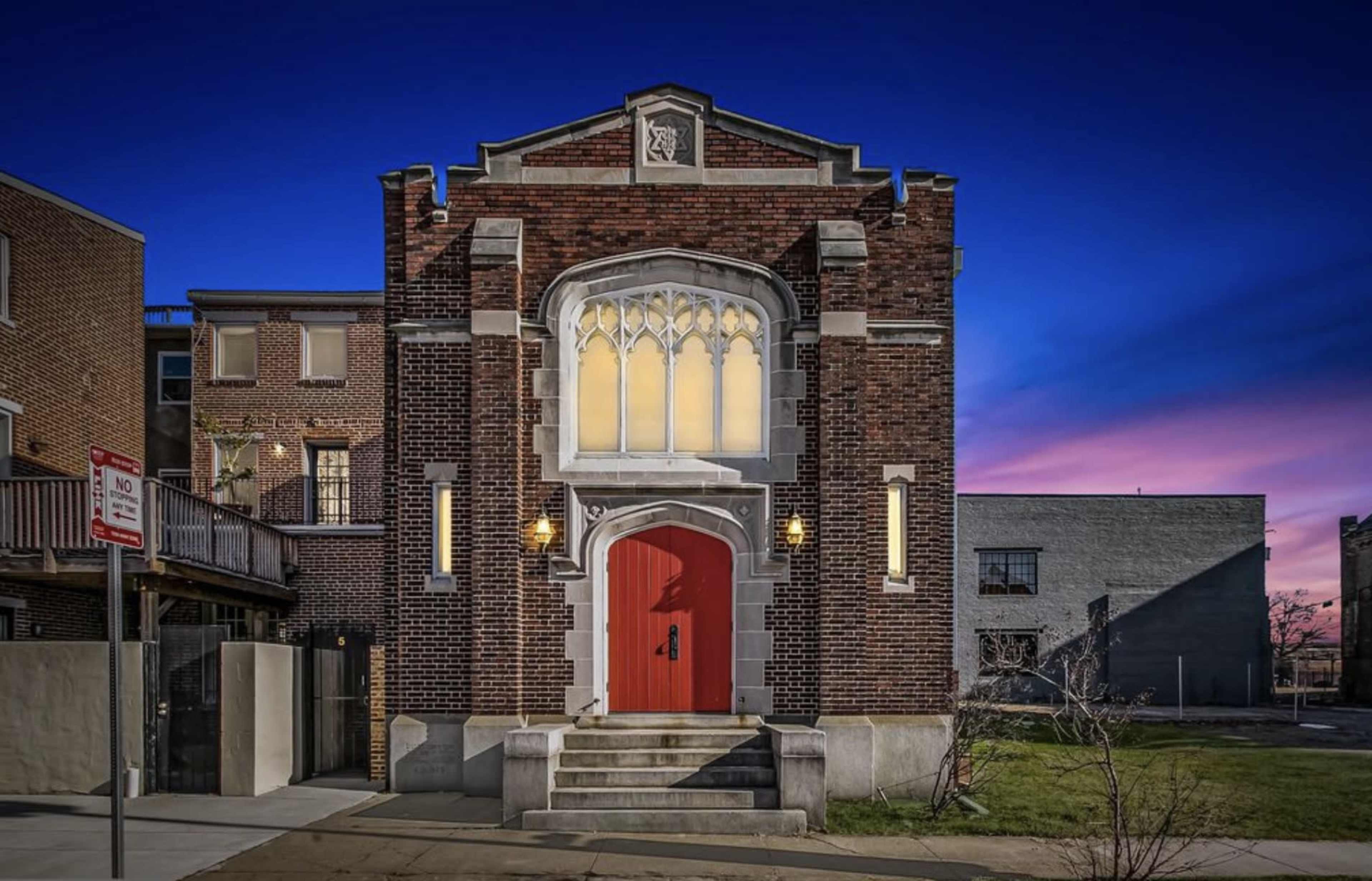 A brick building with a prominent red door and Gothic-style windows stands against a twilight sky.