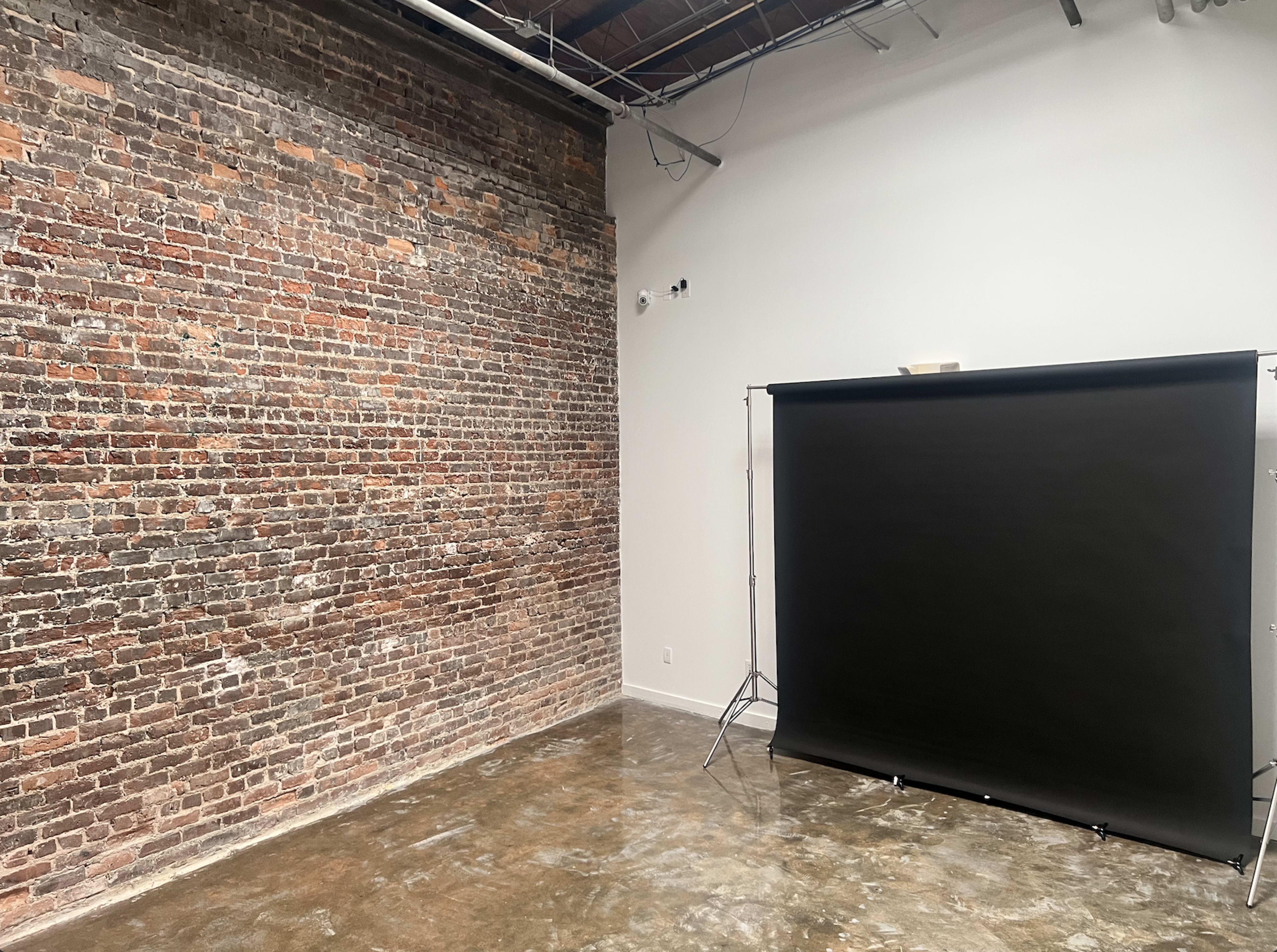 The image shows a spacious room with a rustic brick wall on one side and a black backdrop set up on a stand.