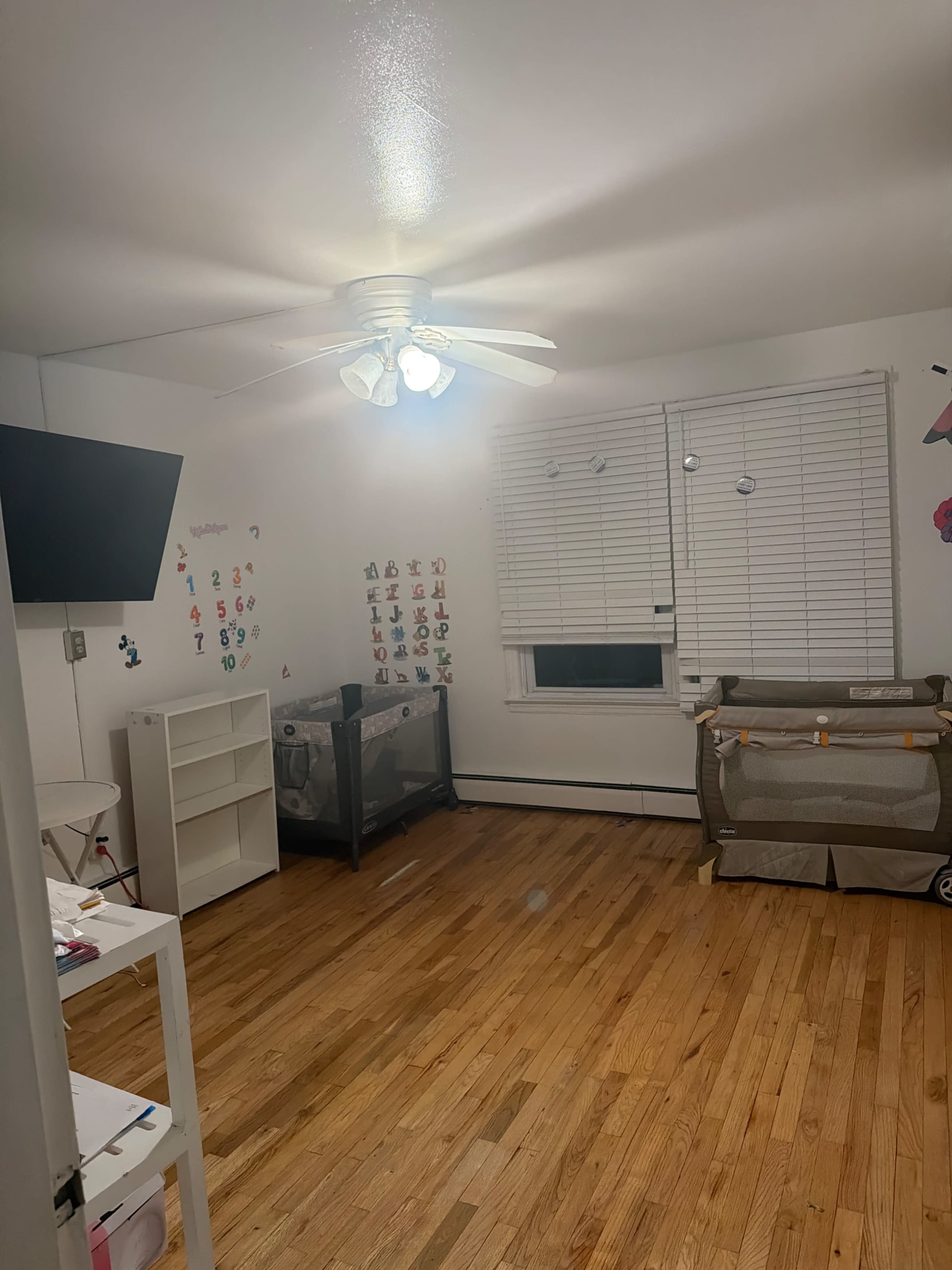 The image shows a sparsely furnished child's room with a crib, a shelving unit, a television on the wall, and light wooden flooring.