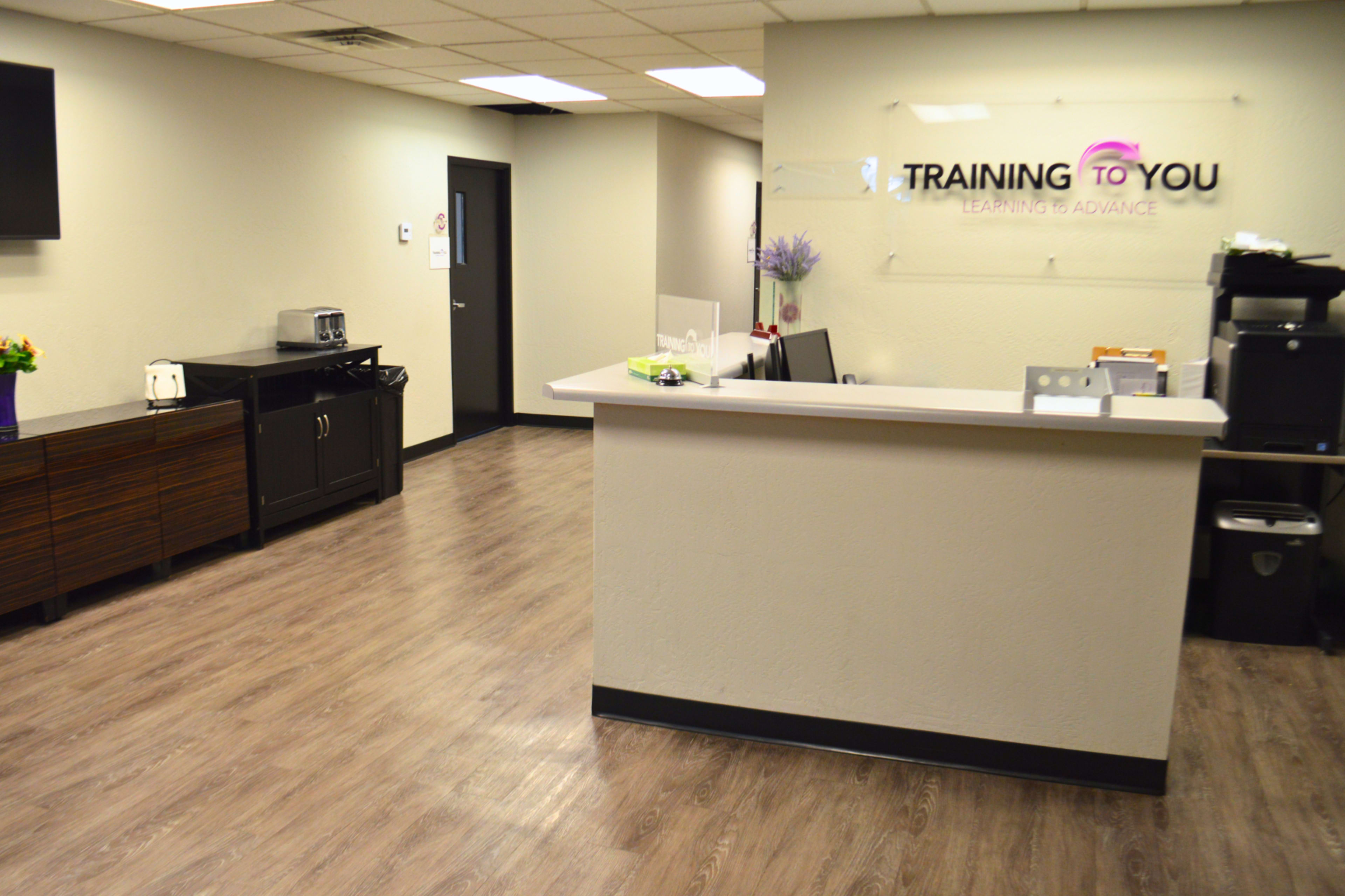 A clean, modern reception area with a counter, kitchenette, and office equipment, featuring a sign that reads "TRAINING TO YOU."